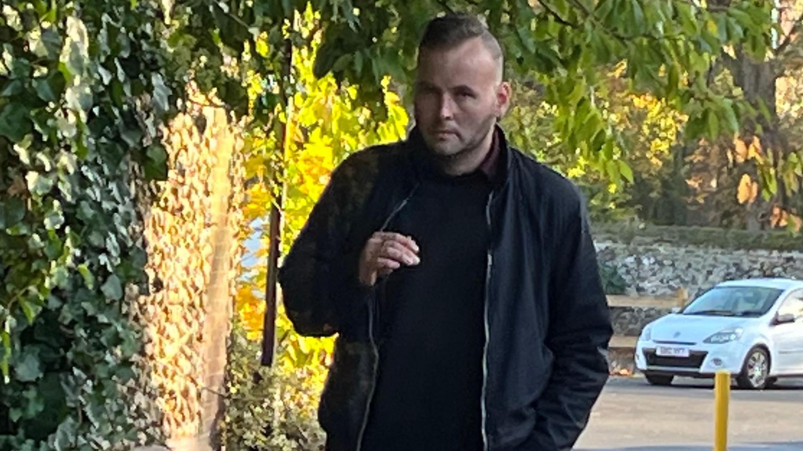 Wayne Finney arriving at Norwich Crown Court wearing all a black jumper and black jacket. He is looking at the camera, unsmiling. In the background you can see a white car. 