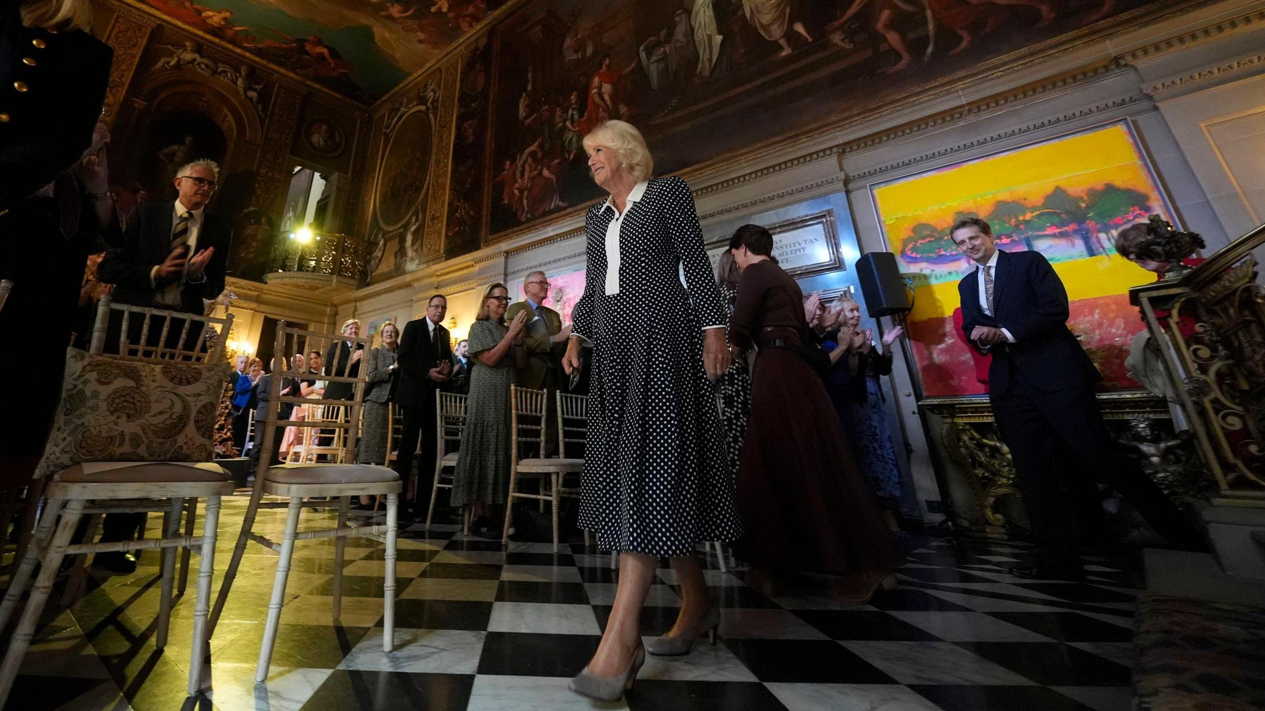 The Queen walking into the Painted Hall where people are on their feet applauding her entrance.