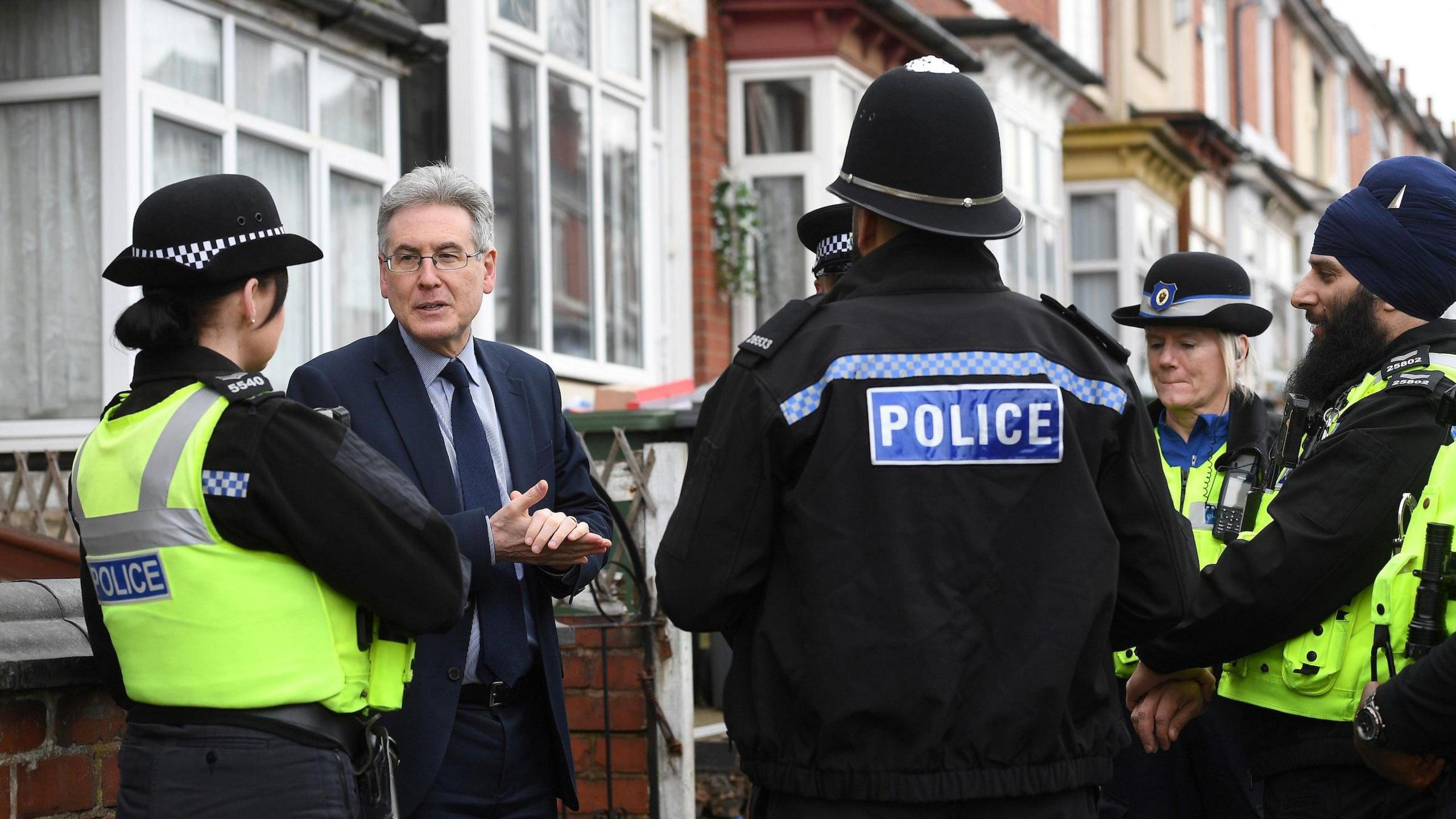 A group of police officers gathered on a residential street. They are standing in a circle and talking to a man in a navy suit and tie. The man is the West Midlands Police and Crime Commissioner. He is standing with his hands together and is in mid-conversation. The officers are all in uniform and some have their backs to the camera.