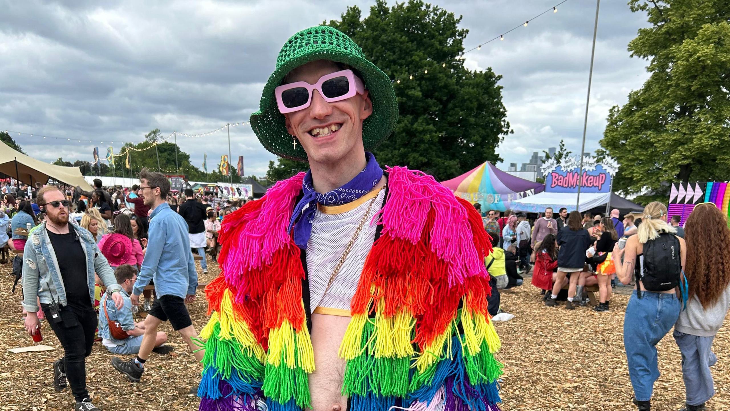 A man smiles at the camera at a music festival. He is wearing a green crocheted bowler hat, pink thick-rimmed sunglasses and a purple bandana around his neck. He's also wearing a mesh cropped tanktop and a rainbow coloured jacket covered in different coloured tassels. People can be seen in the background enjoying the festival.