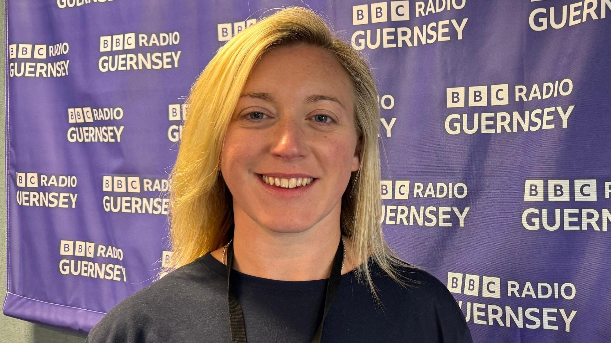 Charlie Cox is smiling at the camera as she stands next to a BBC Radio Guernsey branded banner. She has shoulder-length blonde hair and blue eyes., She is wearing a black top and a lanyard.