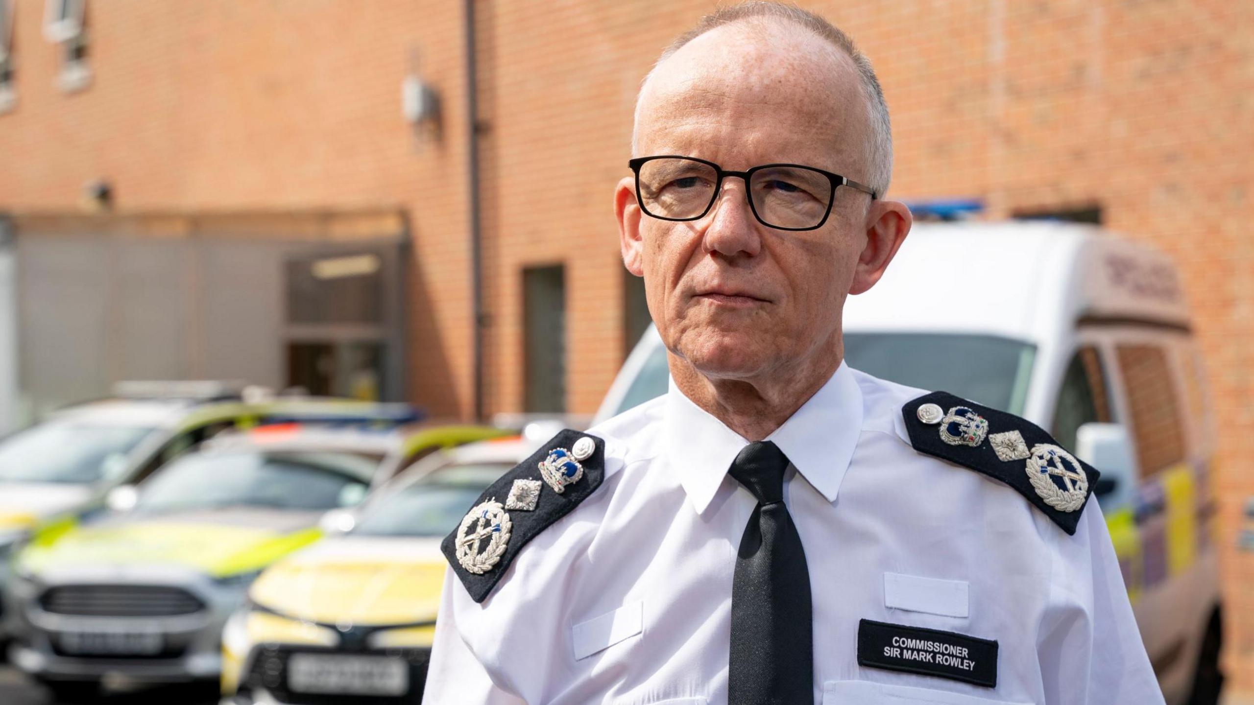 Sir Mark Rowley, an older white man with dark-framed glasses and hair cropped short, wearing a police uniform with with the commissioner's rank insignia on the epaulettes. He is standing in what appears to be the yard of a police station, with a police van and three police cars behind him.