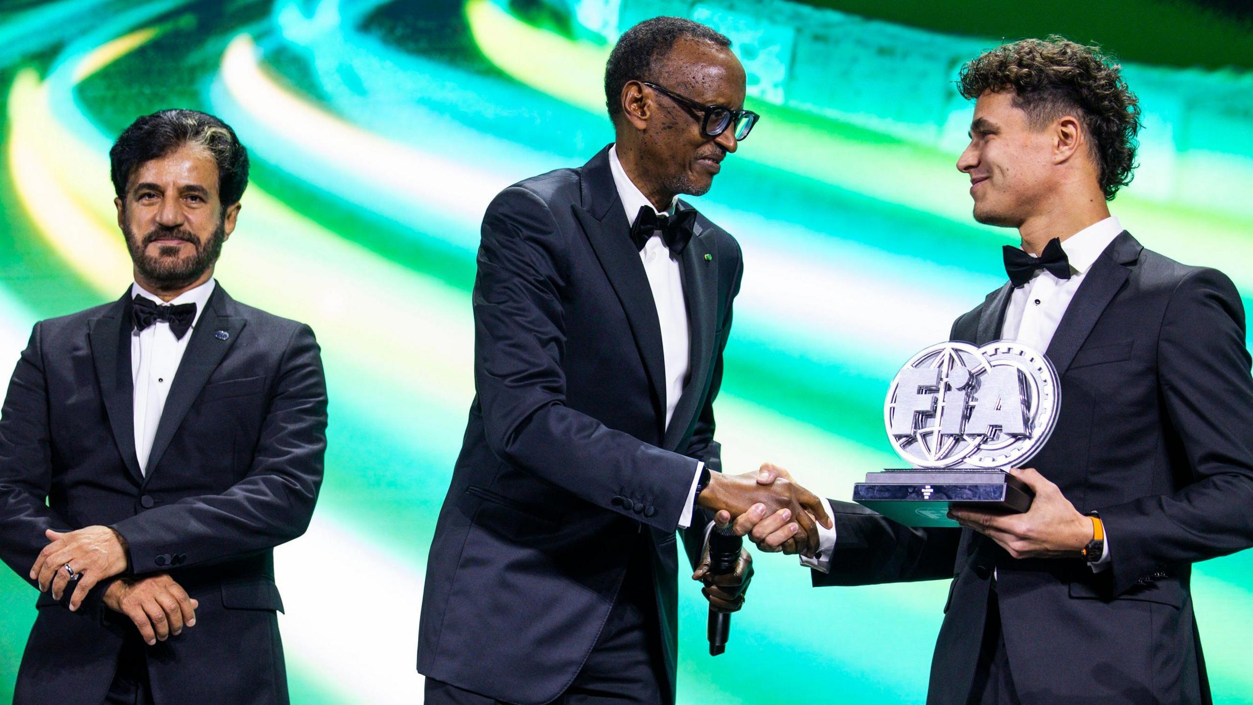 Mohammed Ben Sulayem, President Paul Kagame and Lando Norris are all seen in dinner jackets and bow ties as Kagame and Norris shake hands while the latter holds an FIA trophy