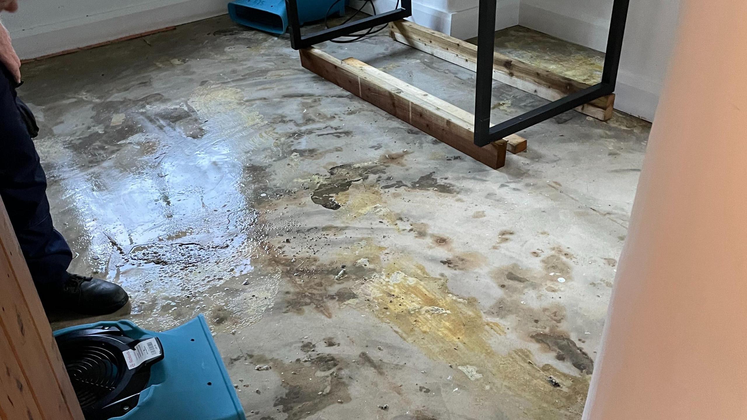 A floor is shown to be stained and flooded with grey/brown water. Wooden furniture is seen in the corner. The reflection of puddles can be seen.