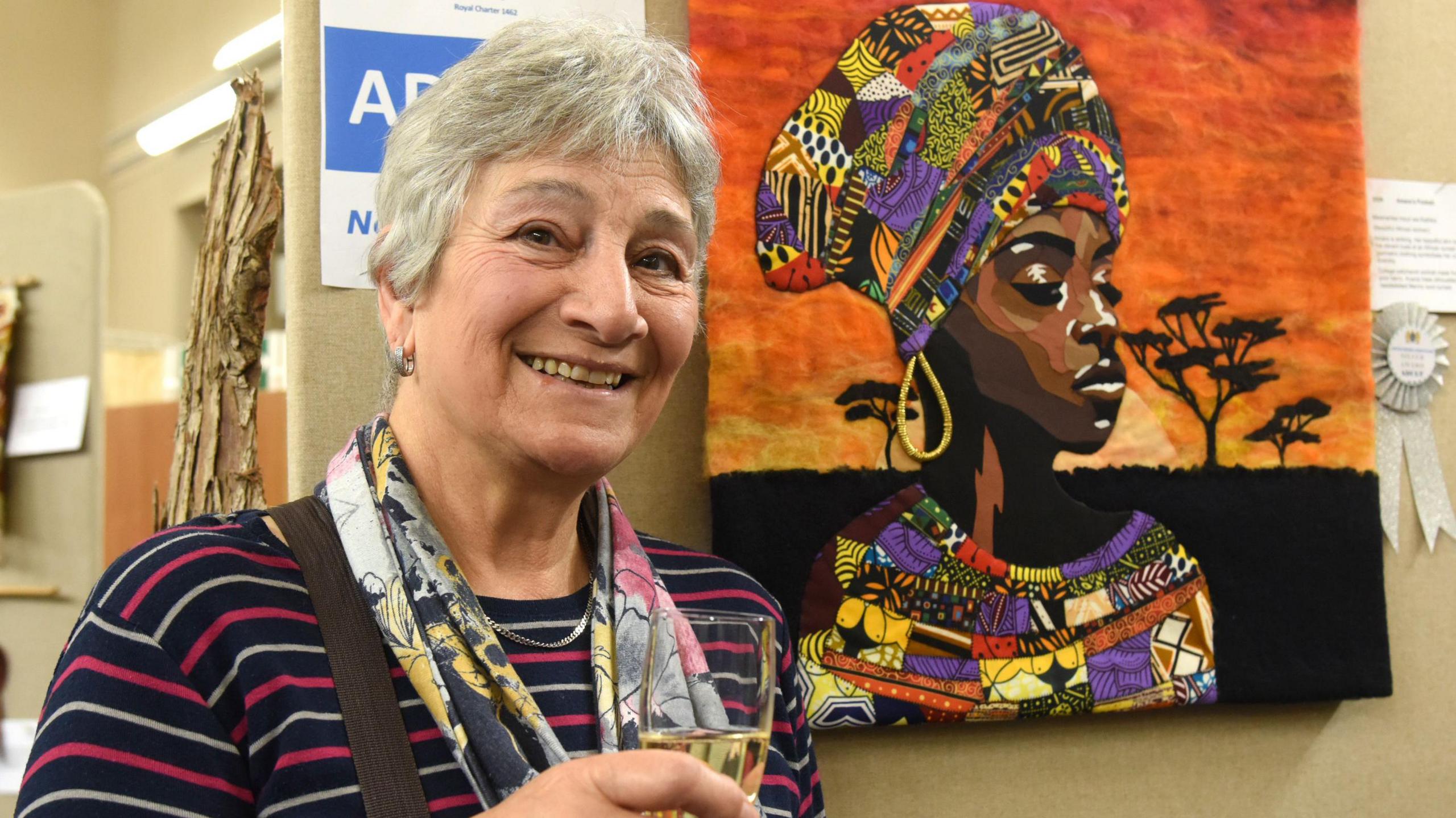 Karen Wright with short grey hair smiling and holding a glass of champagne next to her picture of an African woman made of fabric built up to depict shading on her face and bright colours on her clothes