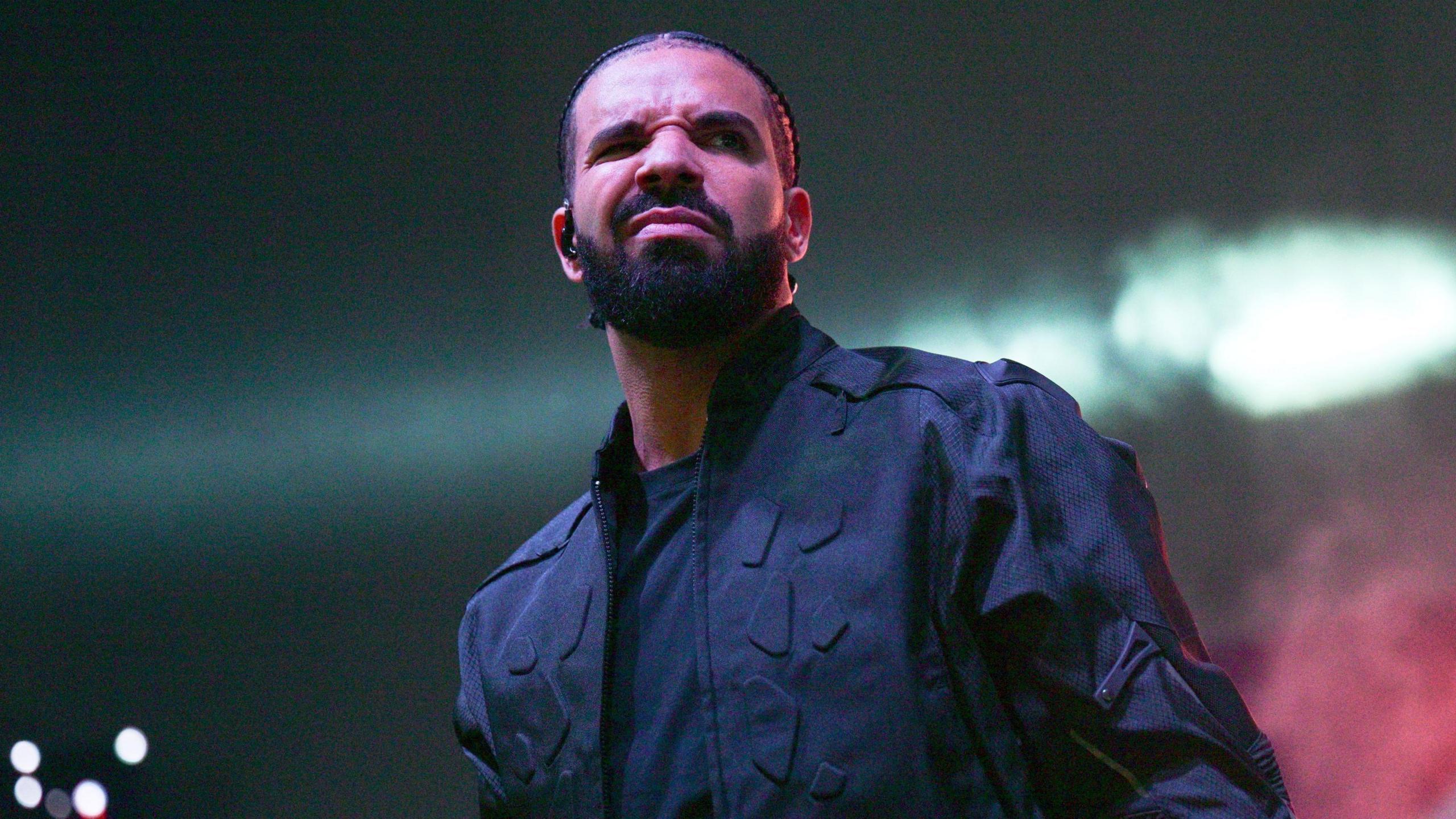 Drake on stage. Drake wears his hair braided and has a short beard. He squints at the camera, his mouth tightly shut. He wears a black jacket over black T-shirt. 