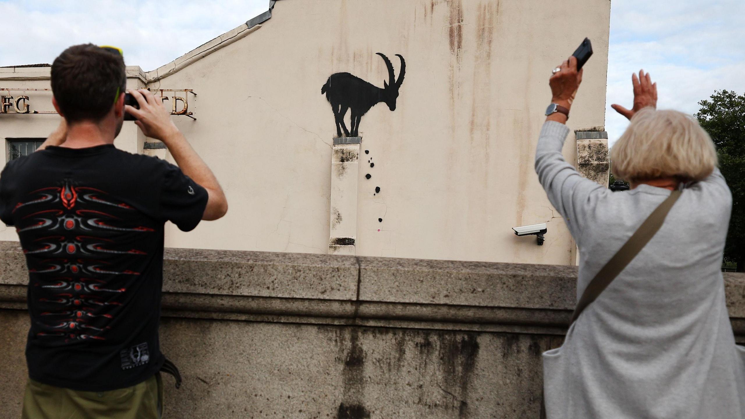 A man and a woman with smartphones in their hands taking pictures of piece of art by street artist Banksy. The work itself is of a goat and it is on the side of a building in Kew in south-west London. 