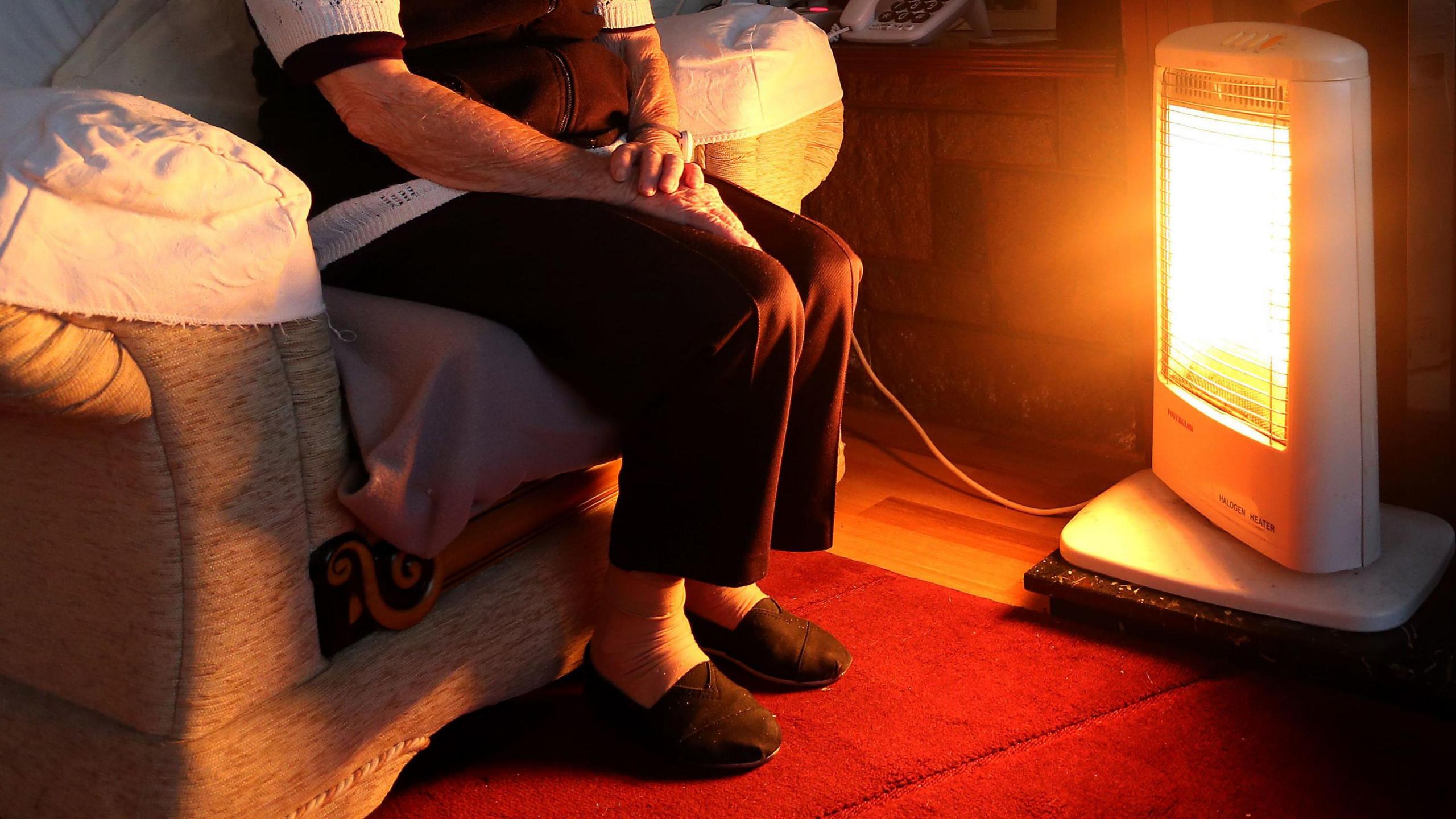 An elderly person wearing slippers, dark trousers and a black and white top sits next to a bar heater. The bar heater is on and is glowing orange. The person's face is not visible and they are sitting on a light coloured chair with a white cover. Their feet are on a red rug.