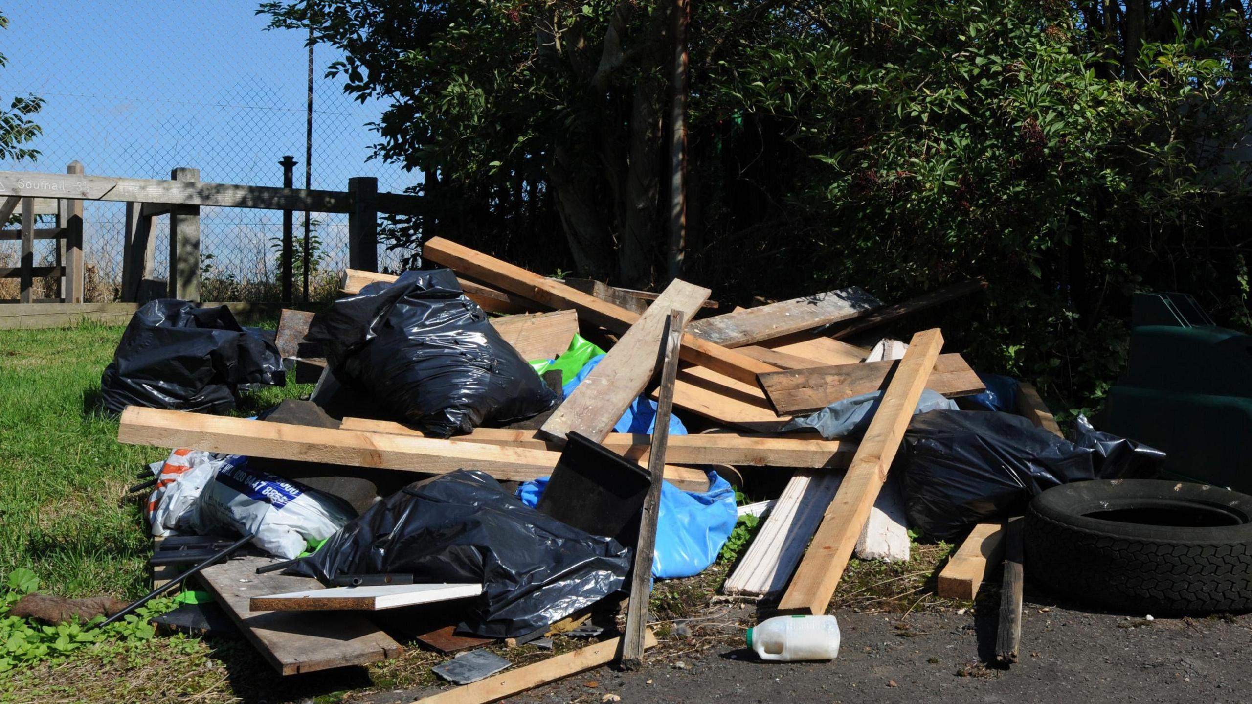 A large pile of fly-tipped waste is on the side of the road. It includes black bin bag and planks of wood as well as random bits of rubbish like a plastic bottle and a tyre. There is a grassy verge and a tree.