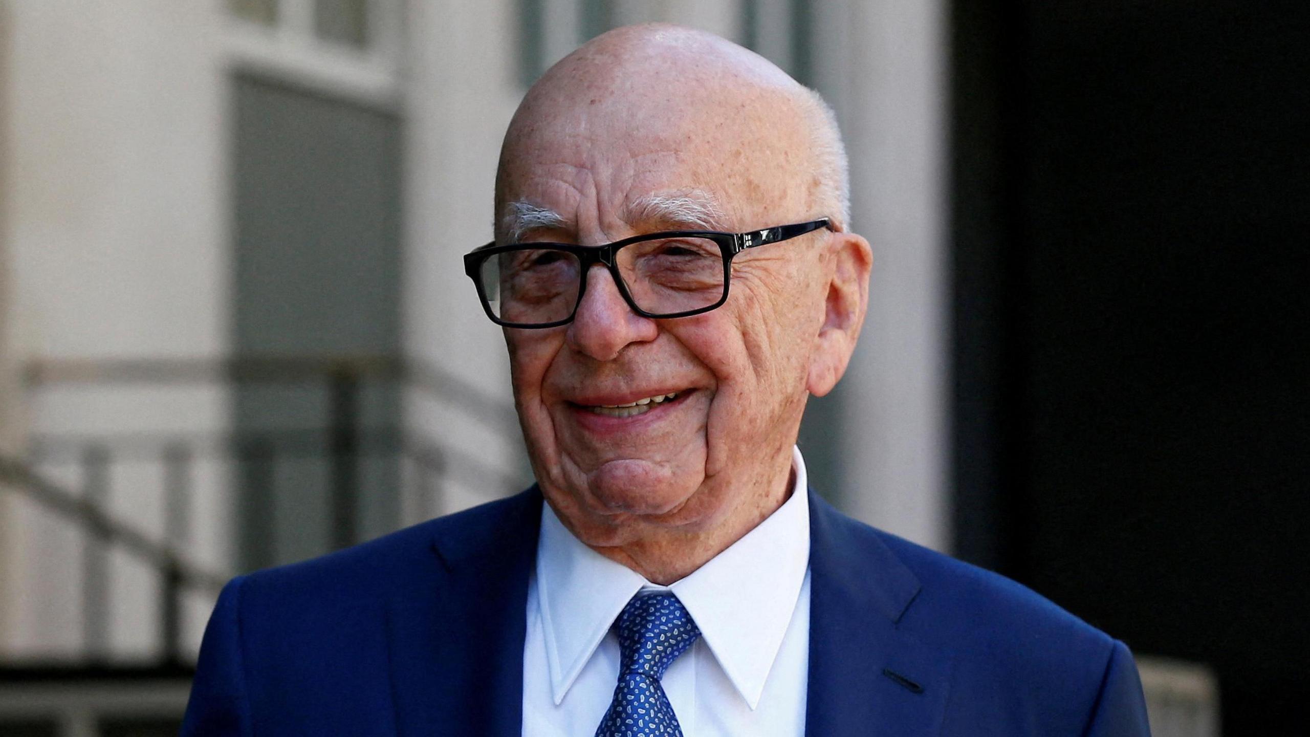 Headshot of Rupert Murdoch standing outside a building wearing a suit and tie and smiling