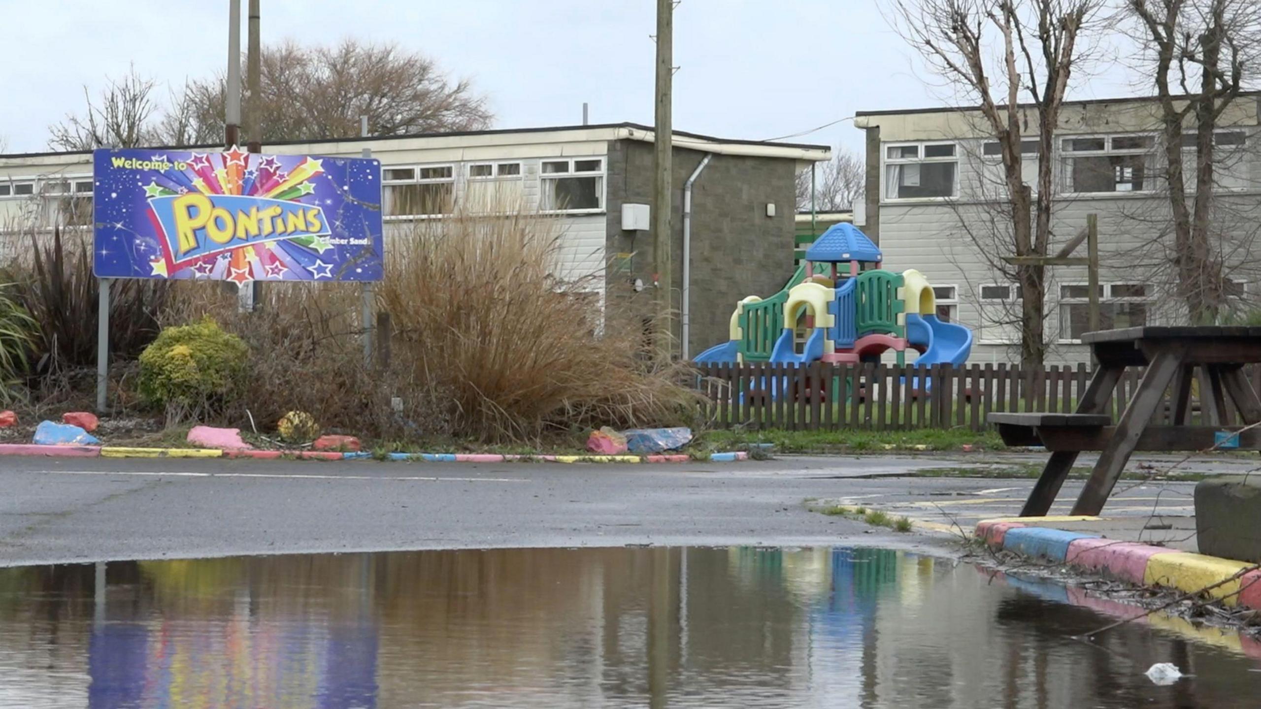 A view of the outside of Pontins holiday park which has been closed for just over a year. There's a large puddle in the foreground. Behind that is some empty holiday holiday chalets and a children's playground. We see a large colourful sign saying "Welcome to Pontins" set in an island of plants which look overgrown and unkempt. 