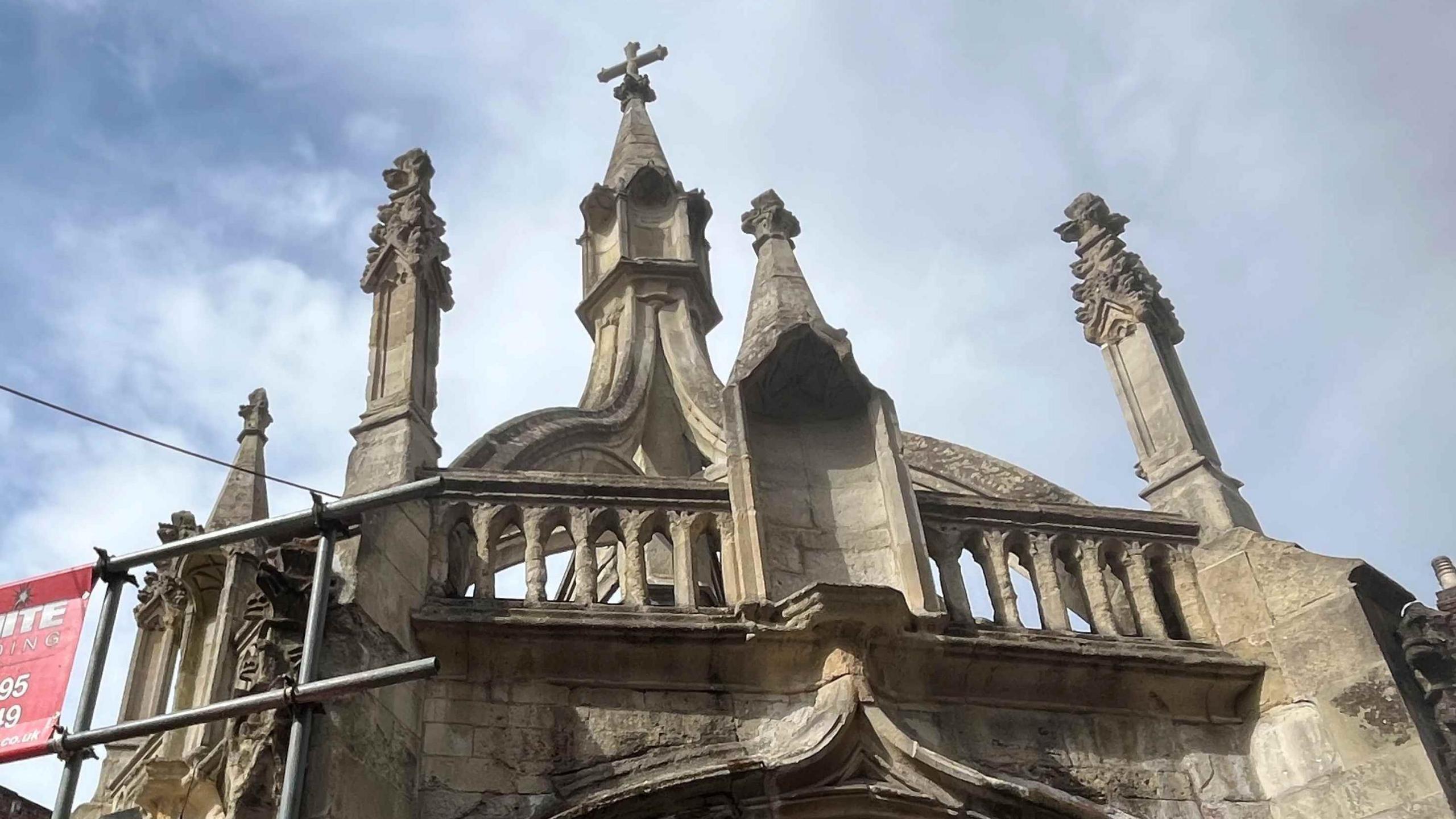 The Poultry Cross. It is a stone, intricately carved structure and there is scaffolding to the left of the building.