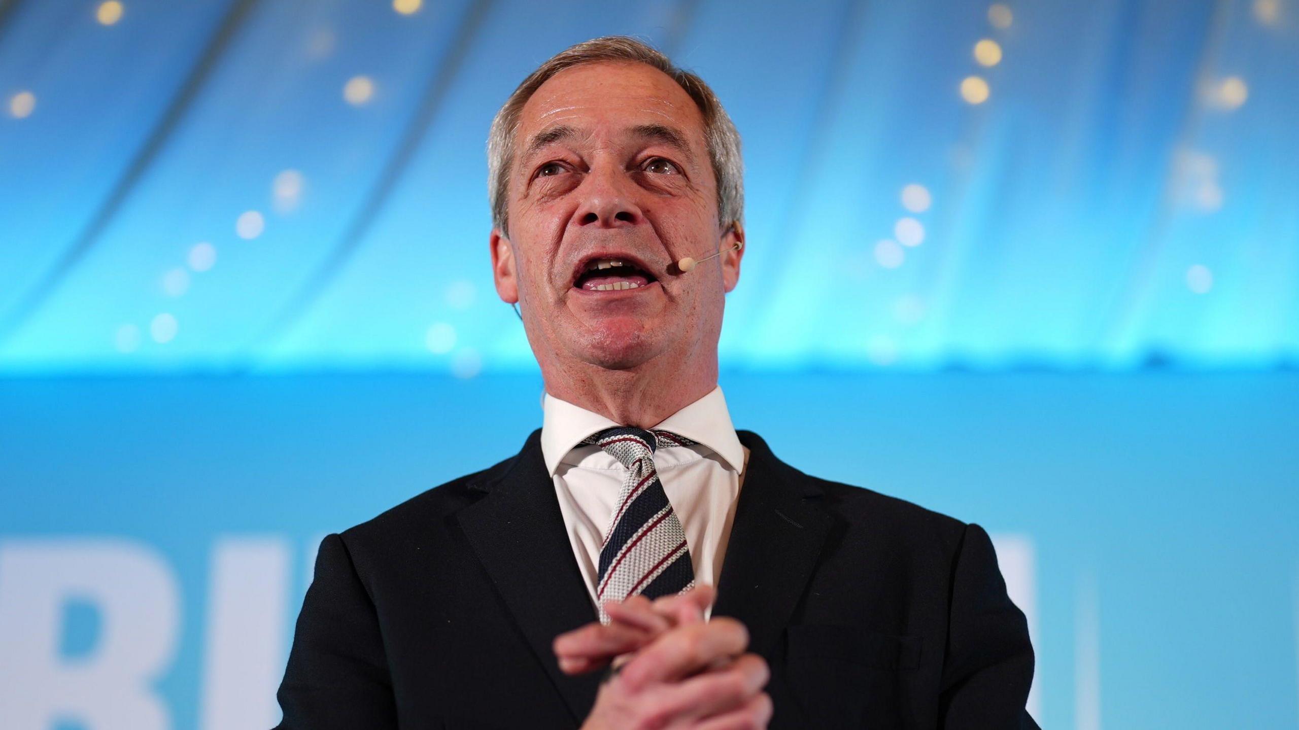 Nigel Farage giving a speech at a rally in North West Essex on Friday night.  he is wearing a dark suit, white shirt and a tie with thick symmetrical stripes which are blue and silver.  The backdrop is in soft focus and the light blue colour of his Reform UK party. 