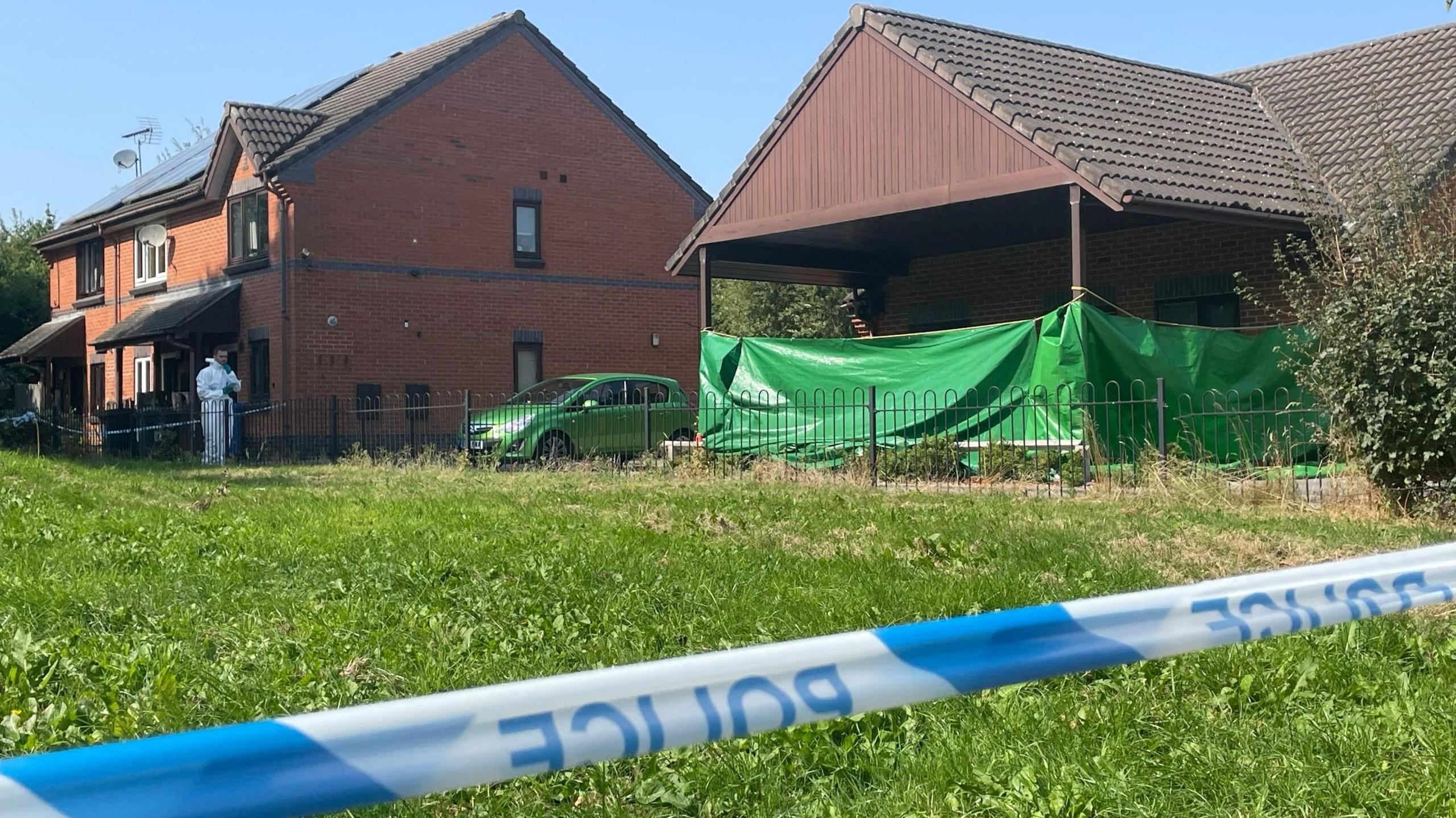 Scene of fire - it shows red brick houses with police tape across grass in front of them. A large section of green tarpaulin is wrapped around of the houses, and a man in a white forensic suit can be seen in the background.