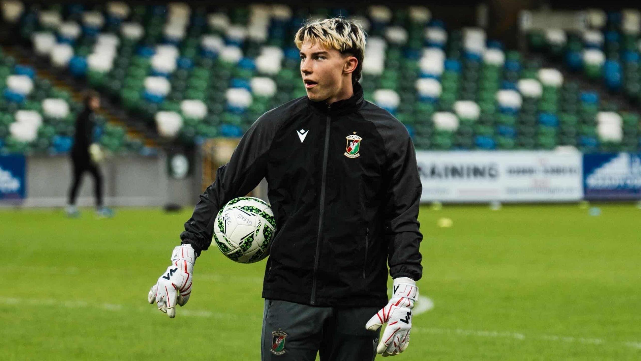 Milo Beimers in football kit including Glentoran training jacket and goalkeeper gloves, on the pitch at a stadium, with a ball under his right arm