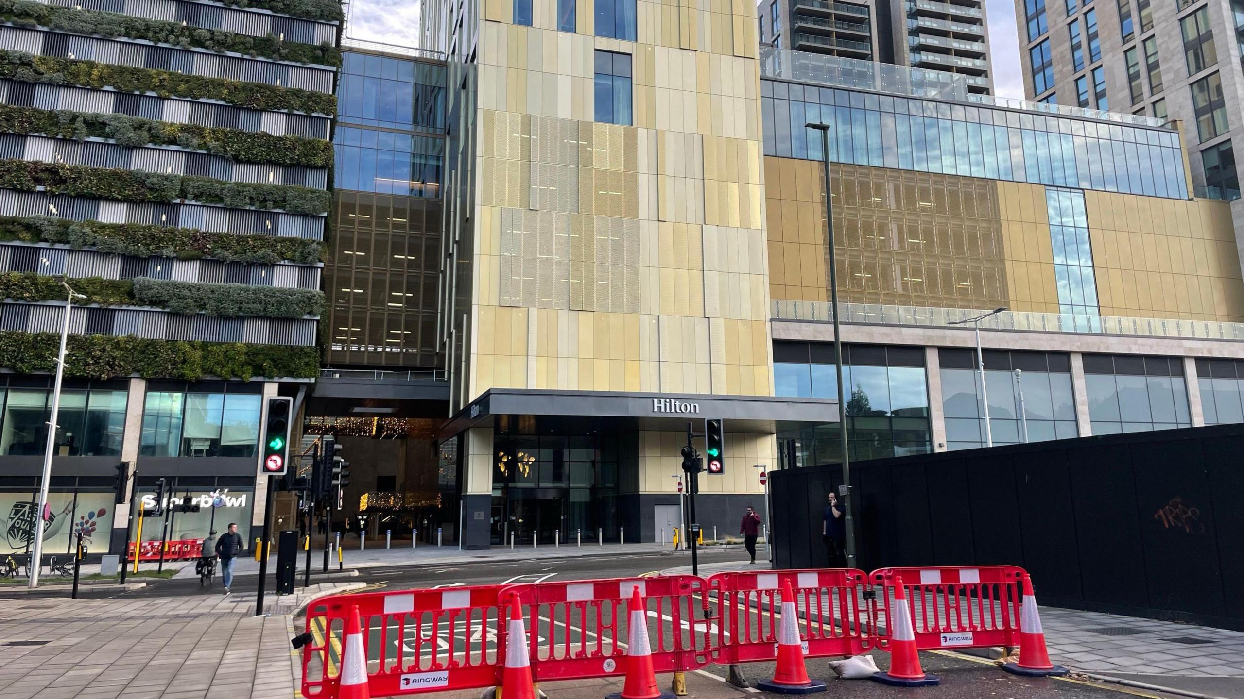 A large hotel with yellow and grey cladding. To the right is a building with plants on the front. In the foreground is a road blocked with orange and white cones and barriers