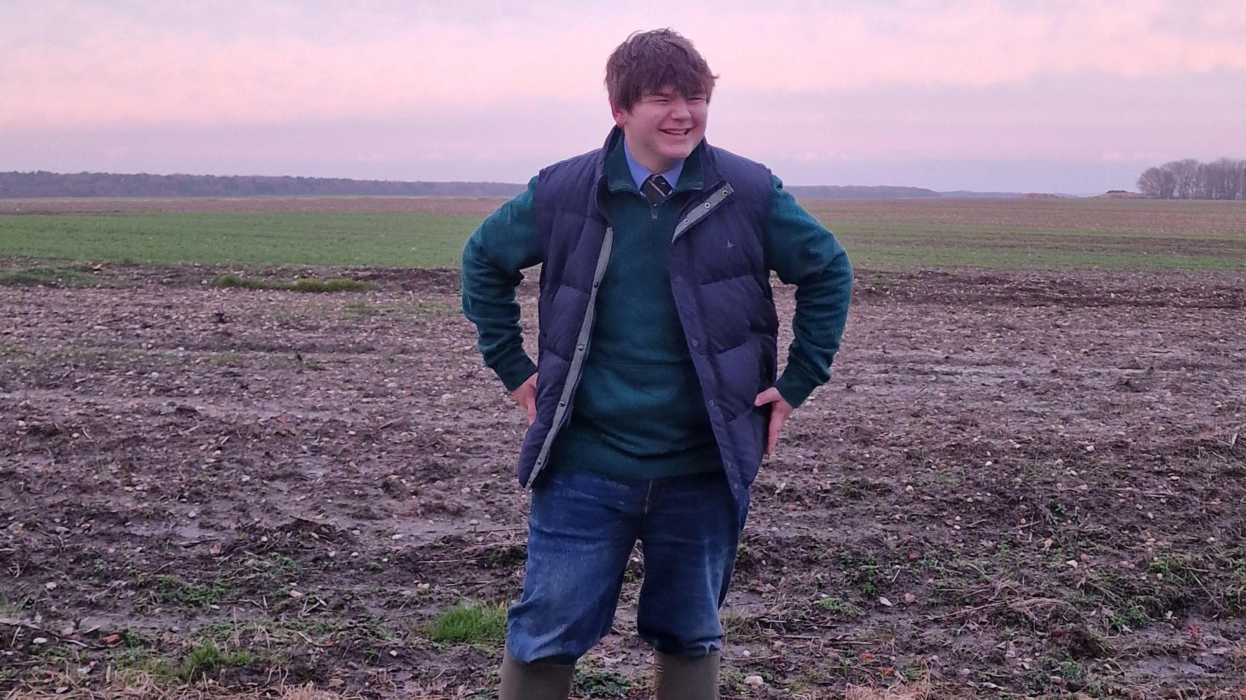A pink sunset can be seen creating a hue across a photo. In the foreground a teenager with brown hair and a fringe can be seen laughing and looking to the side. He is wearing a navy waistcoat, blue fleece with a school tie and shirt underneath. He also has jeans and wellies on and is standing in a field.
