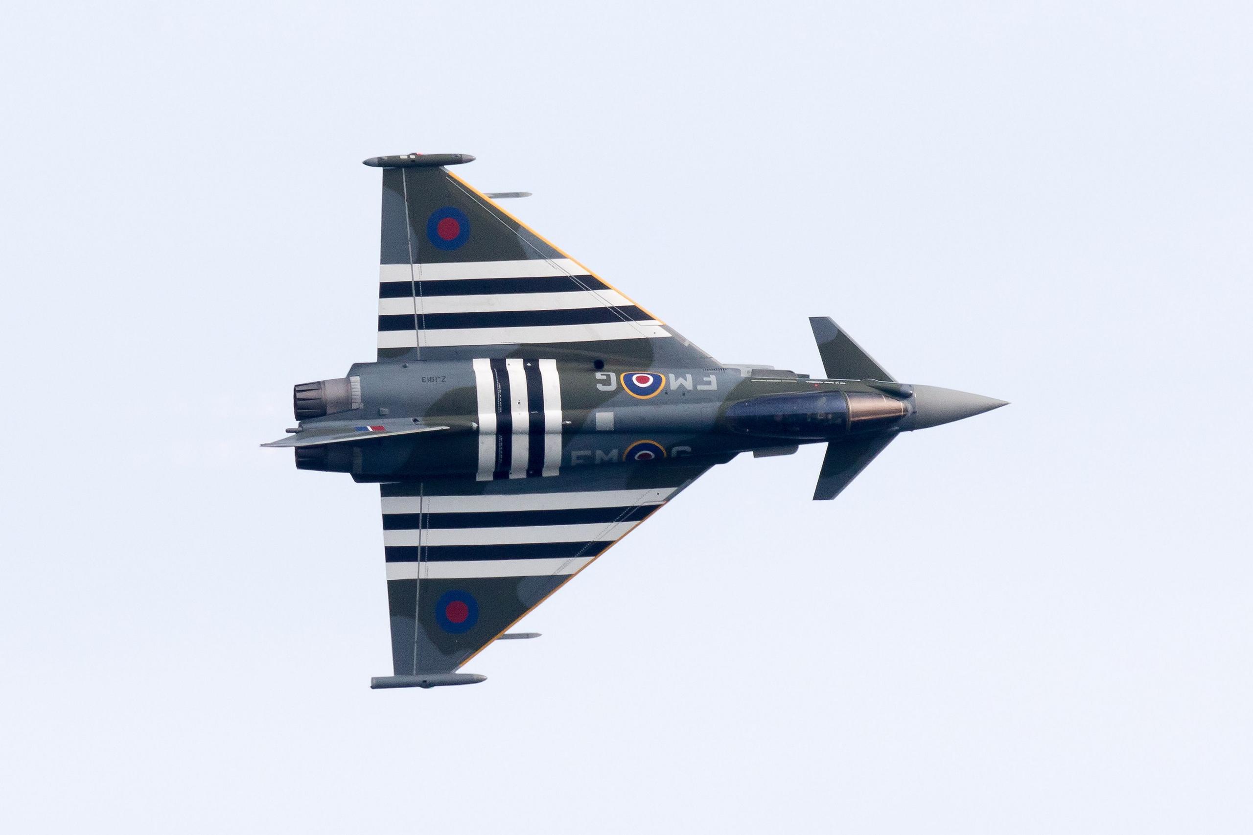 A view of the top of a Typhoon fighter jet as it turns in the sky with the top of the cockpit facing the camera. Its wings and fuselage are painted with the Battle of Britain black and white stripes