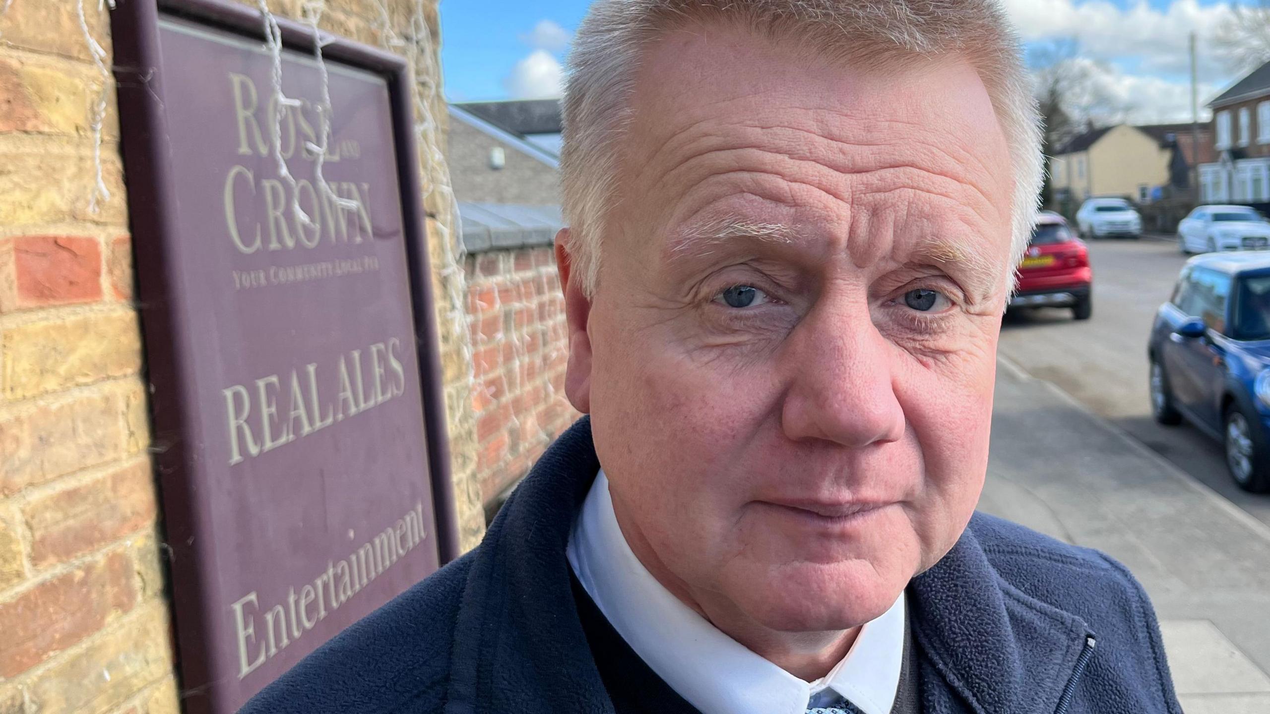 Charlie Marks has reddish short cropped hair and is wearing a blue fleece top over a light blue shirt with a light blue spotty tie. Behind him is a High Street with cars parked on the roadside and housing visible too.