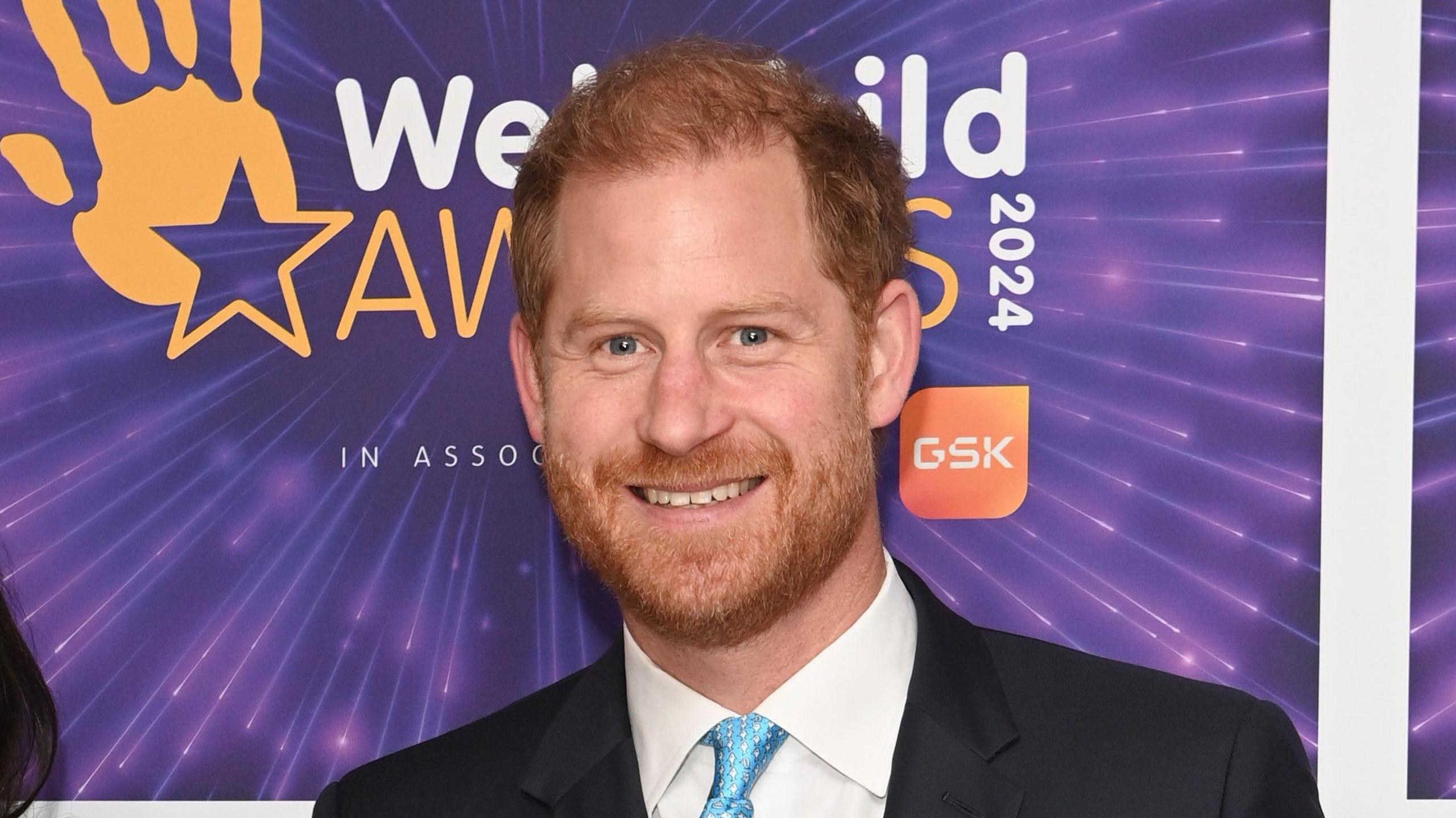 Prince Harry standing in front of a purple background. He is wearing a black suit with a light blue tie.