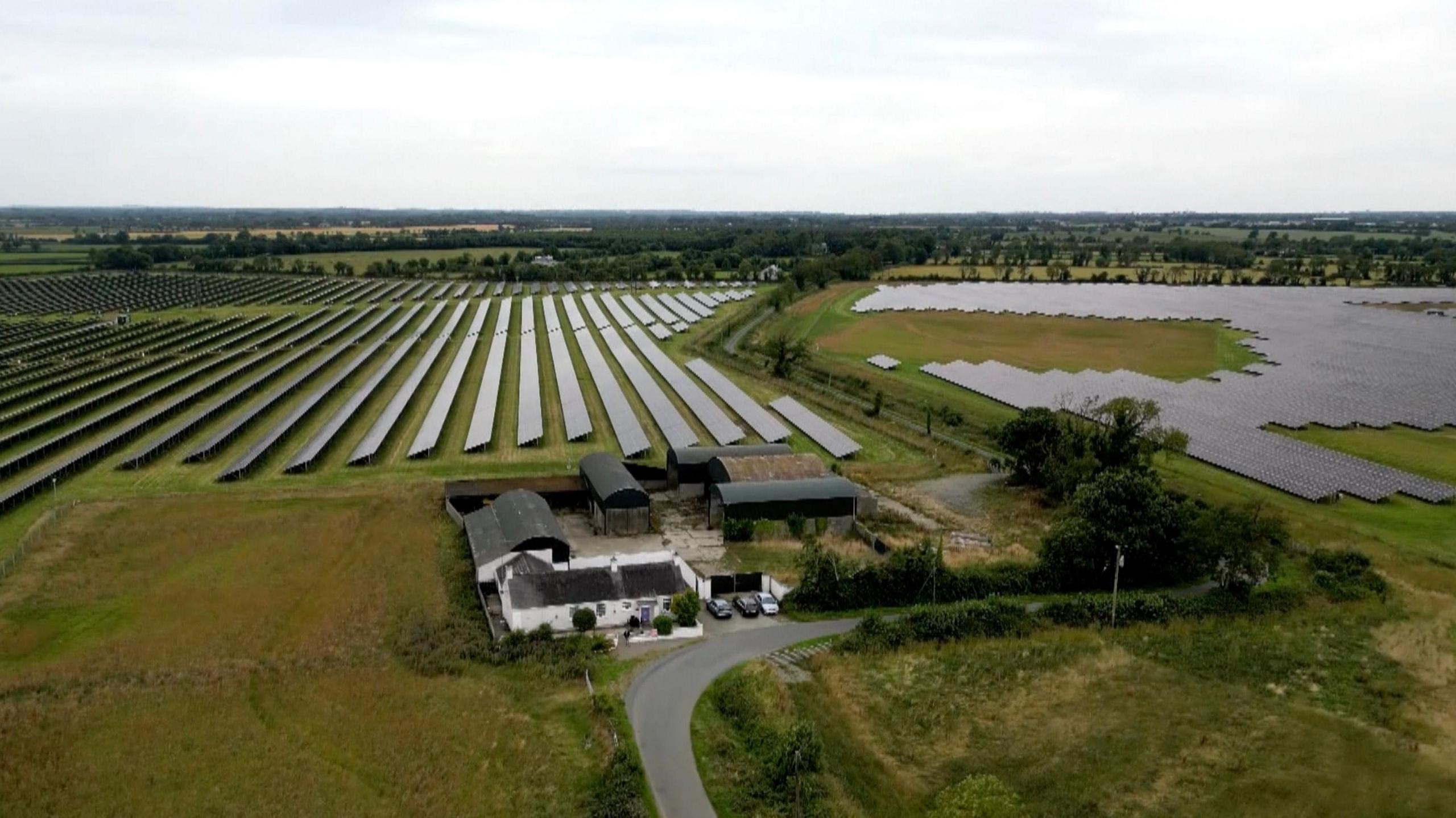 A small farm house and buildings on farm land. It is sounded by trees and large industrial size solar panels.