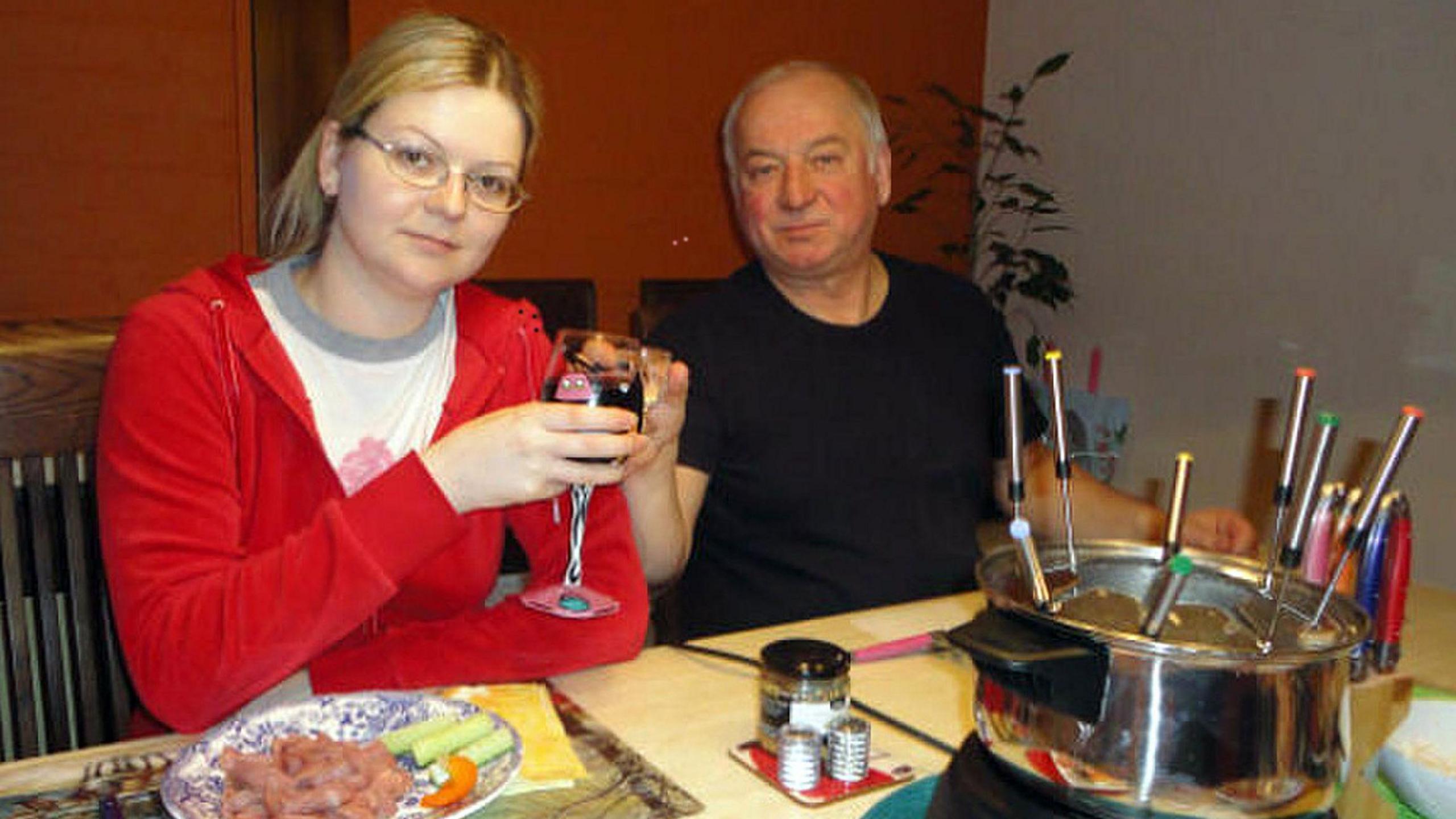 A young woman in a red top clinks a glass with an older man in a black t-shirt while sitting at a table laid with a fondue set. She has a plate of food in front of her. 