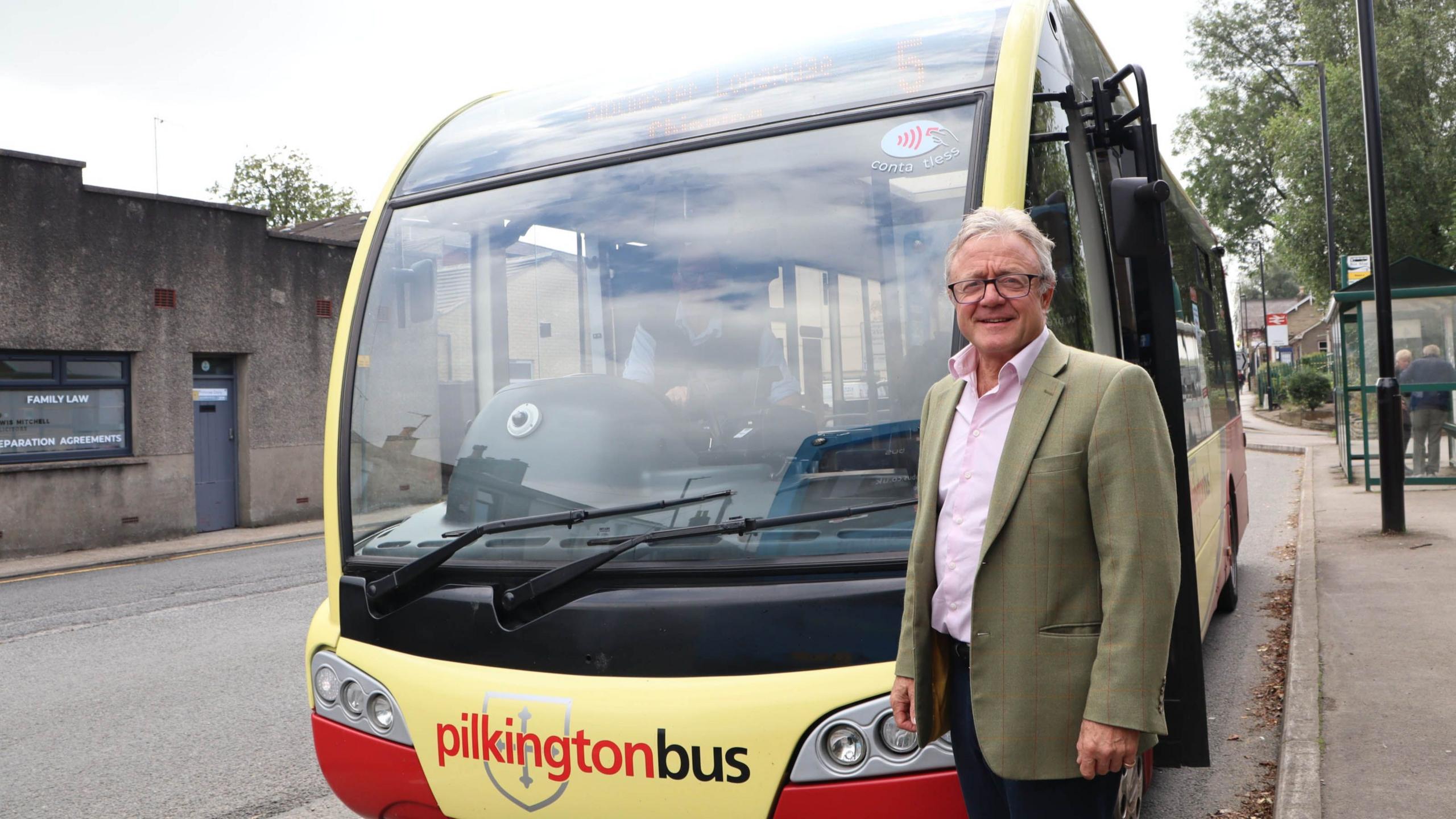 County councillor Rupert Swarbrick standing smiling at a bus stop in front of a yellow and red bus