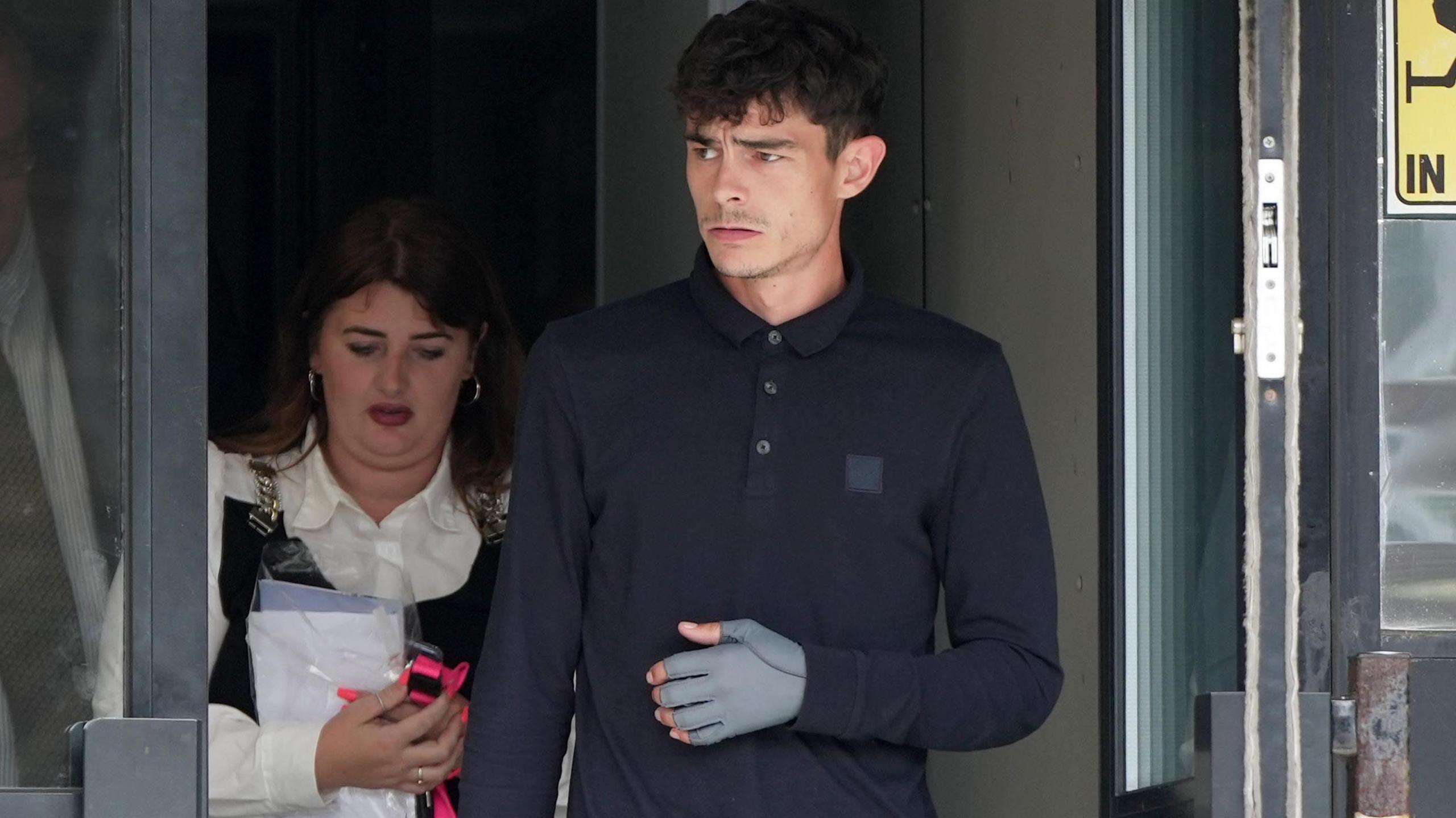 A man with short black hair wearing a dark blue polo shirt. He is walking through a door and looking to the left. Behind him is a woman with long brown hair in a white shirt