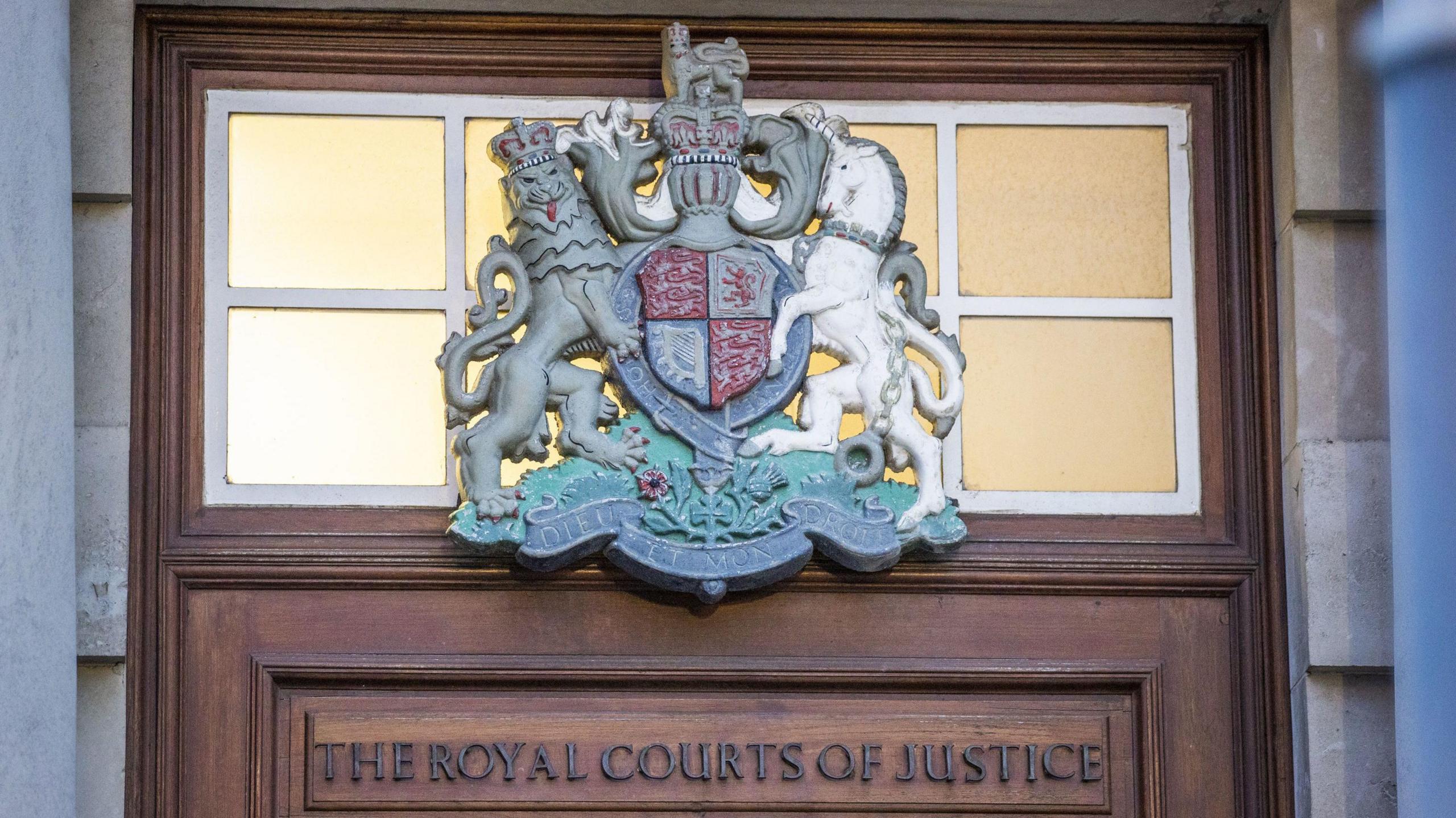 A wooden door frame reads 'The Royal Courts of Justice.' There is a grey and white badge with a lion, on the left, and a white horse on the right facing each other. Between them is a blue and red crest in the shape of a shield. Both animals stand on their hind legs facing the shield. A red crown sits on top of the shield.