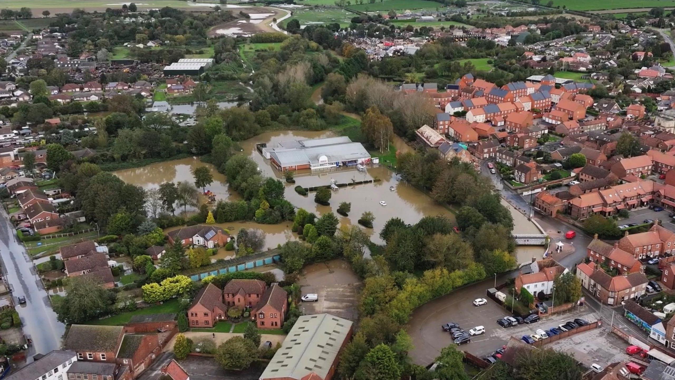 A drone view of a flooded neighbourhood in Horncastle. Brown water covers fields and surrounds buildings, with trees rising above the water.