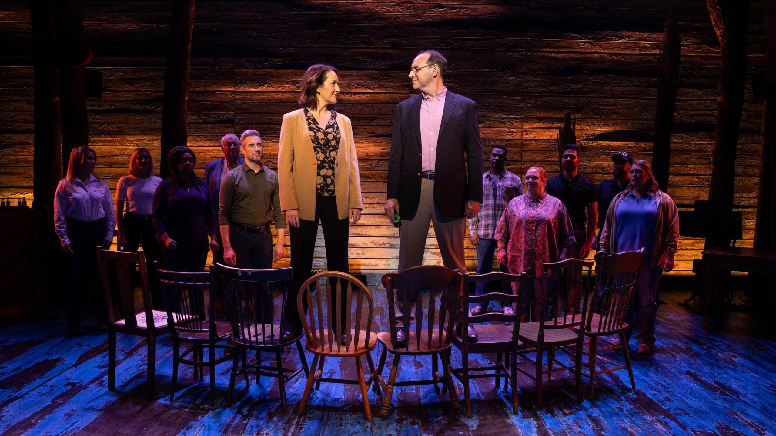 A production shot from a scene in Come From Away. A man and a woman have their heads turned towards one another as they are stood on a line of wooden chairs. A group of people stand behind them, looking at them. The man is dressed in a blazer and chinos, and the woman in black trousers and a sand-coloured jacket. The spotlight is on the couple. 