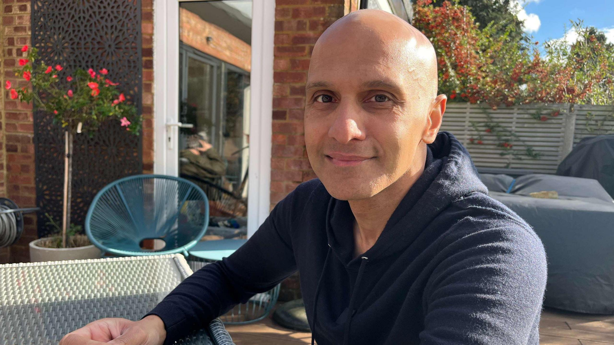 A middle aged man sits in a neat garden. He is bald and is wearing a navy coloured hoodie. Garden furniture and the side of a red brick house are in the background. 