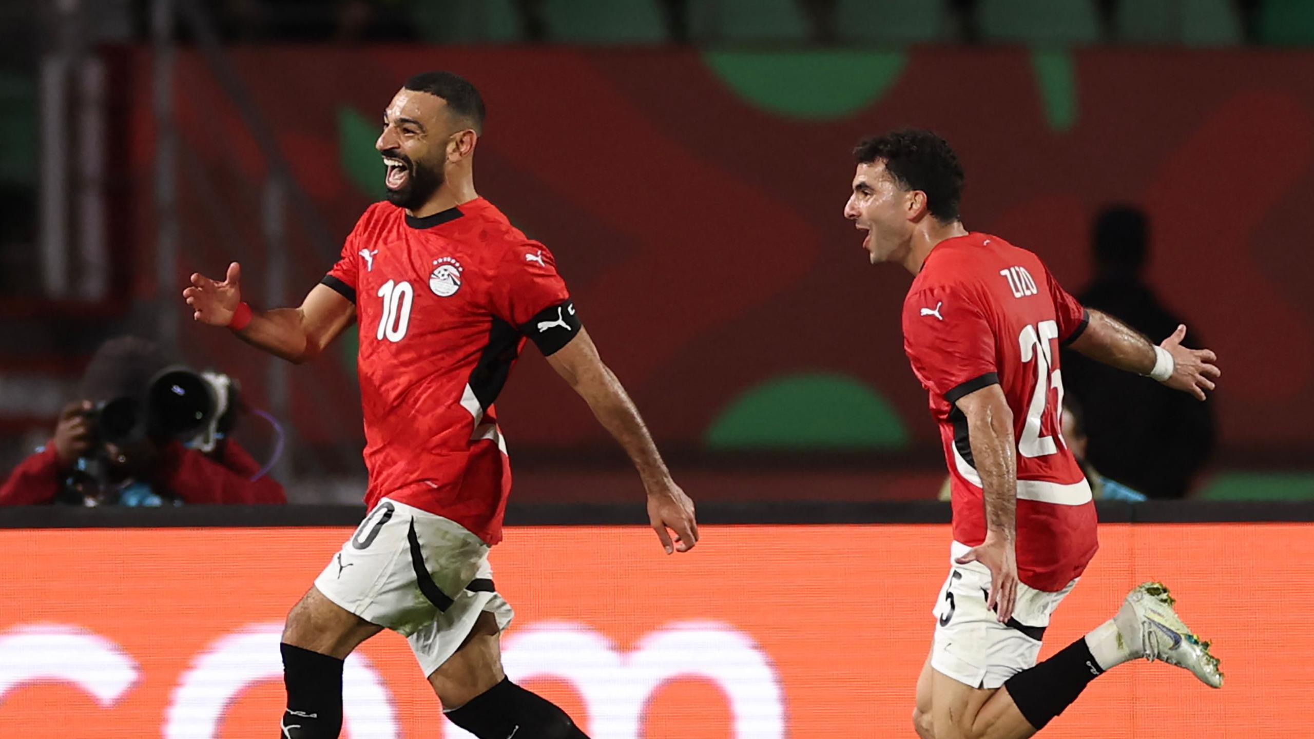 Mohamed Salah (left) celebrates scoring a goal for Egypt with his team-mate Zizo running after him. Both are in red Egypt jerseys, white shorts and black socks and have delighted smiles on their faces and arms outstretched