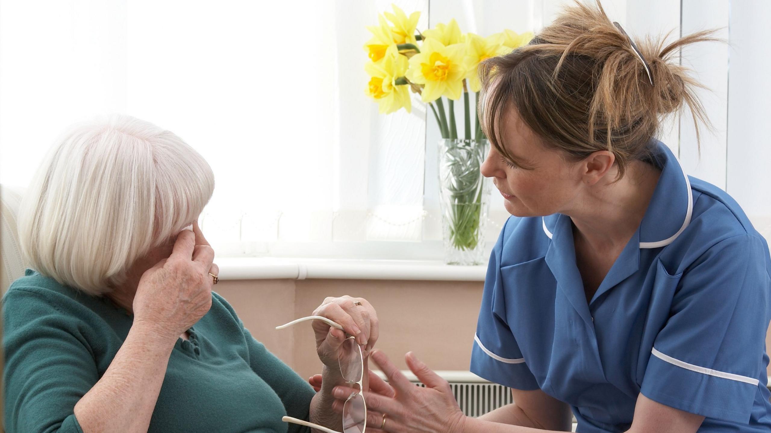A nurse in a blue uniform is comforting an older woman who is wiping her eyes