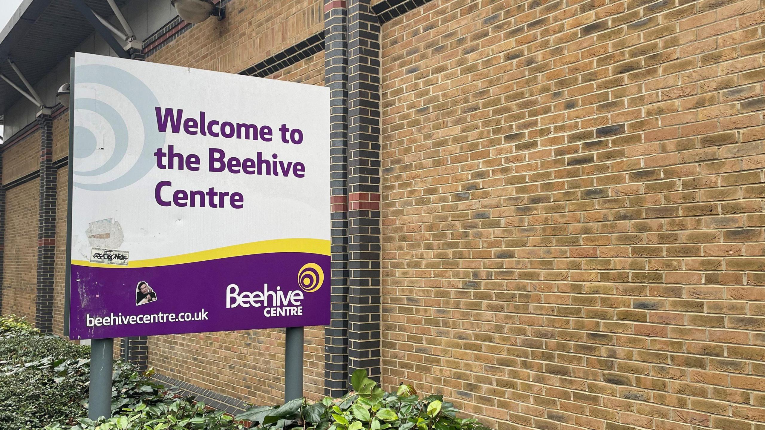 A white, yellow and purple sign next to a brick building says "welcome to the Beehive Centre".