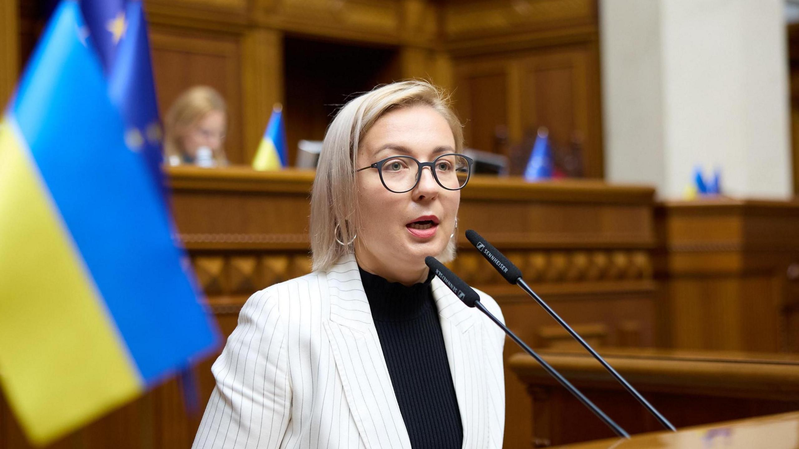 Inna Sovsun speaking in parliament in Kyiv with a Ukrainian flag behind her.