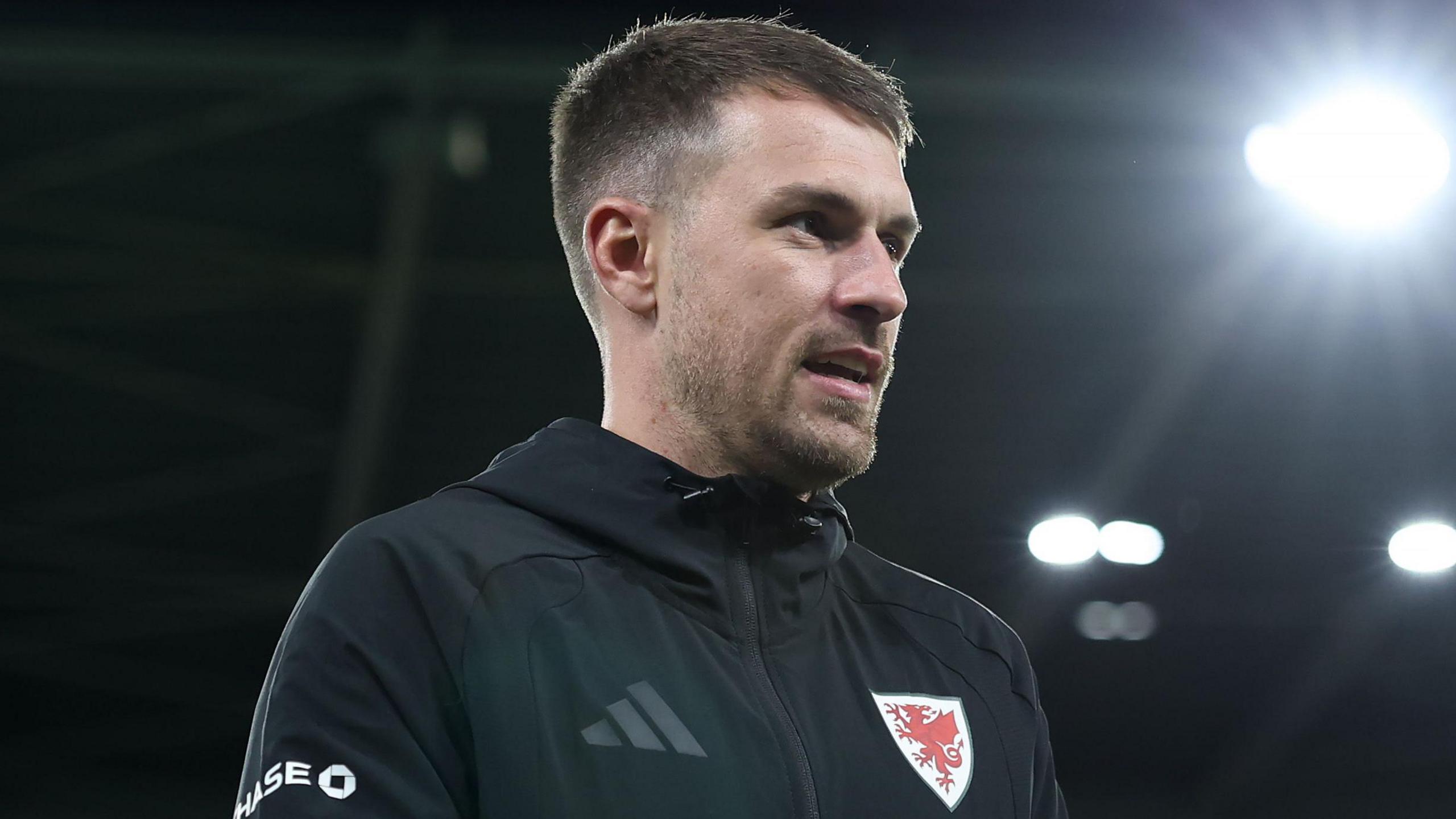 Aaron Ramsey, wearing a black Wales national team tracksuit top with the Welsh crest on the left of his chest, looking on before a game against Macedonia in November 2025.