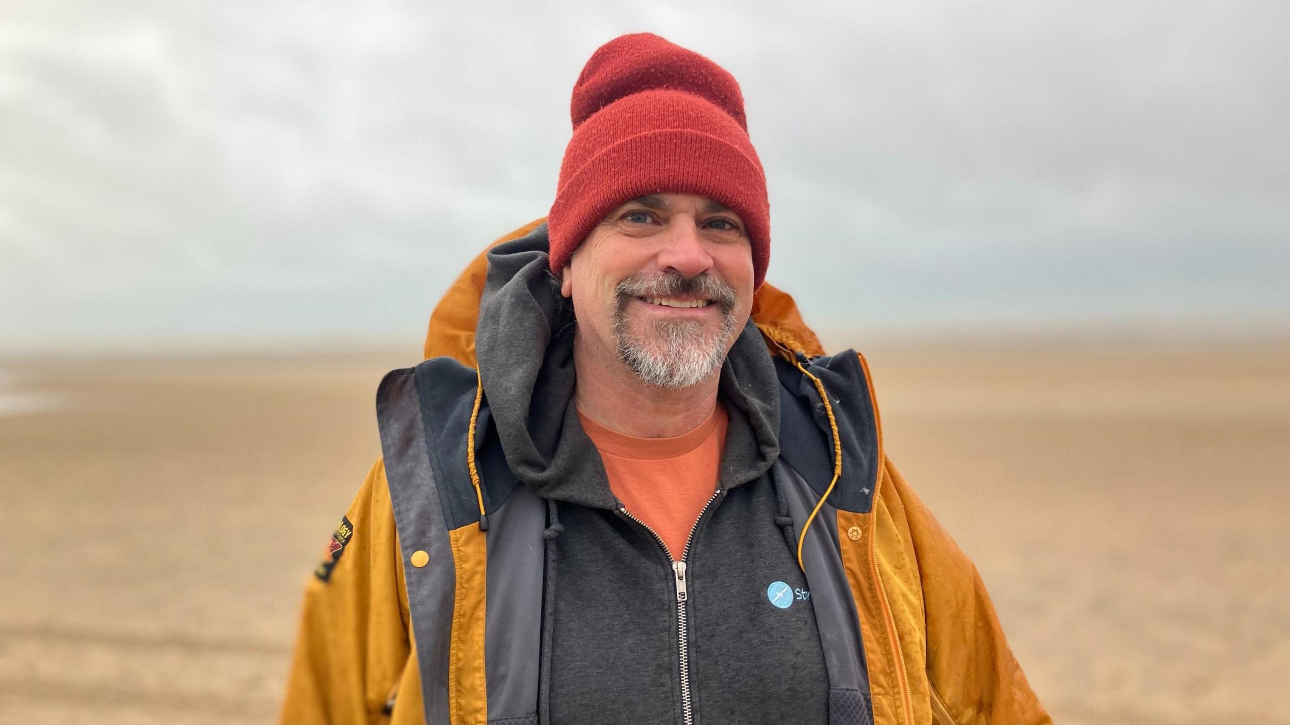 Dominic Manning stands on Camber Sands beach. He wears an orange t-shirt, a grey hoody and a yellow waterproof.