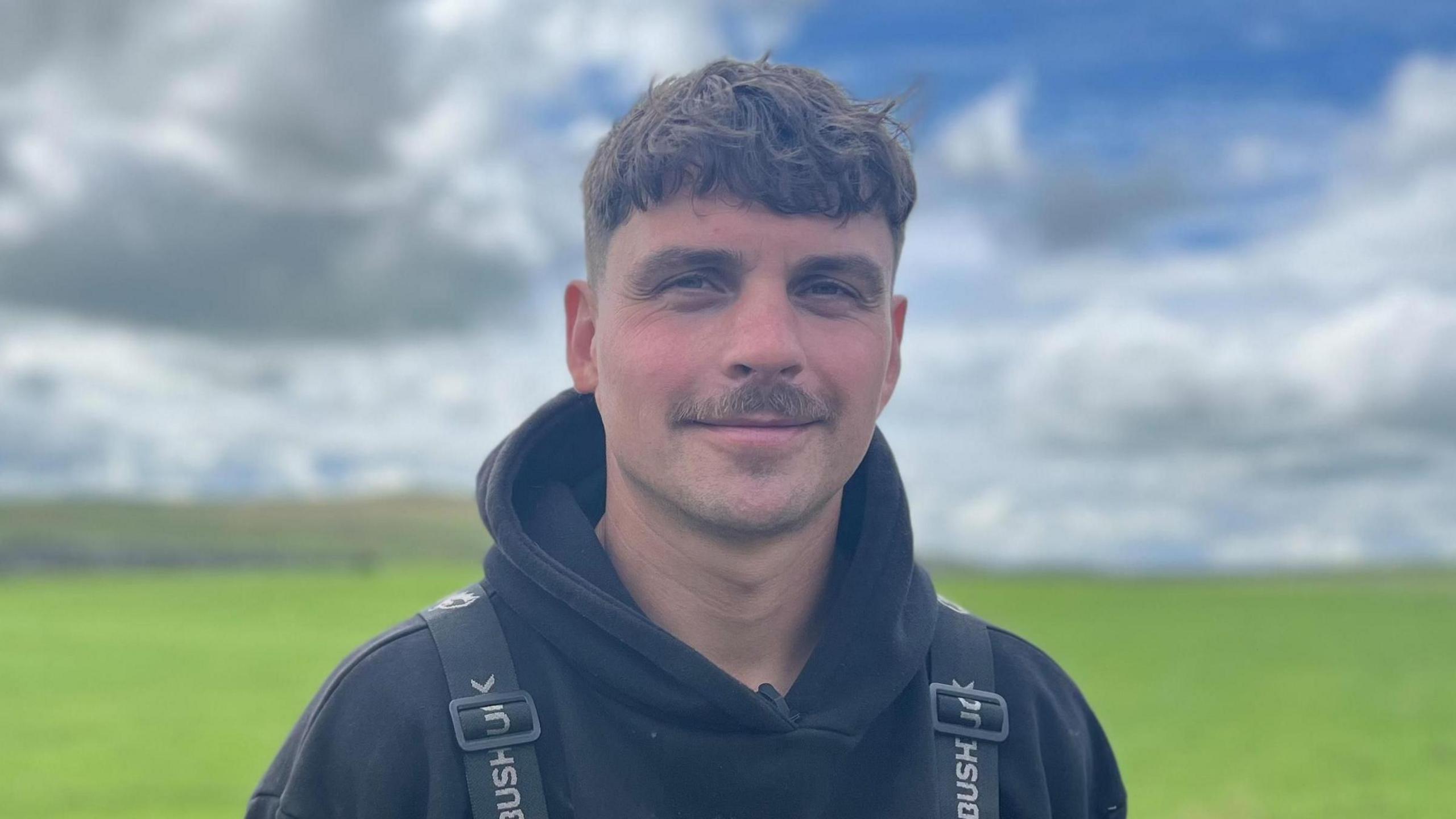 Frank Carr, who has short dark hair and a moustache. He is wearing a black hoody and black overalls.