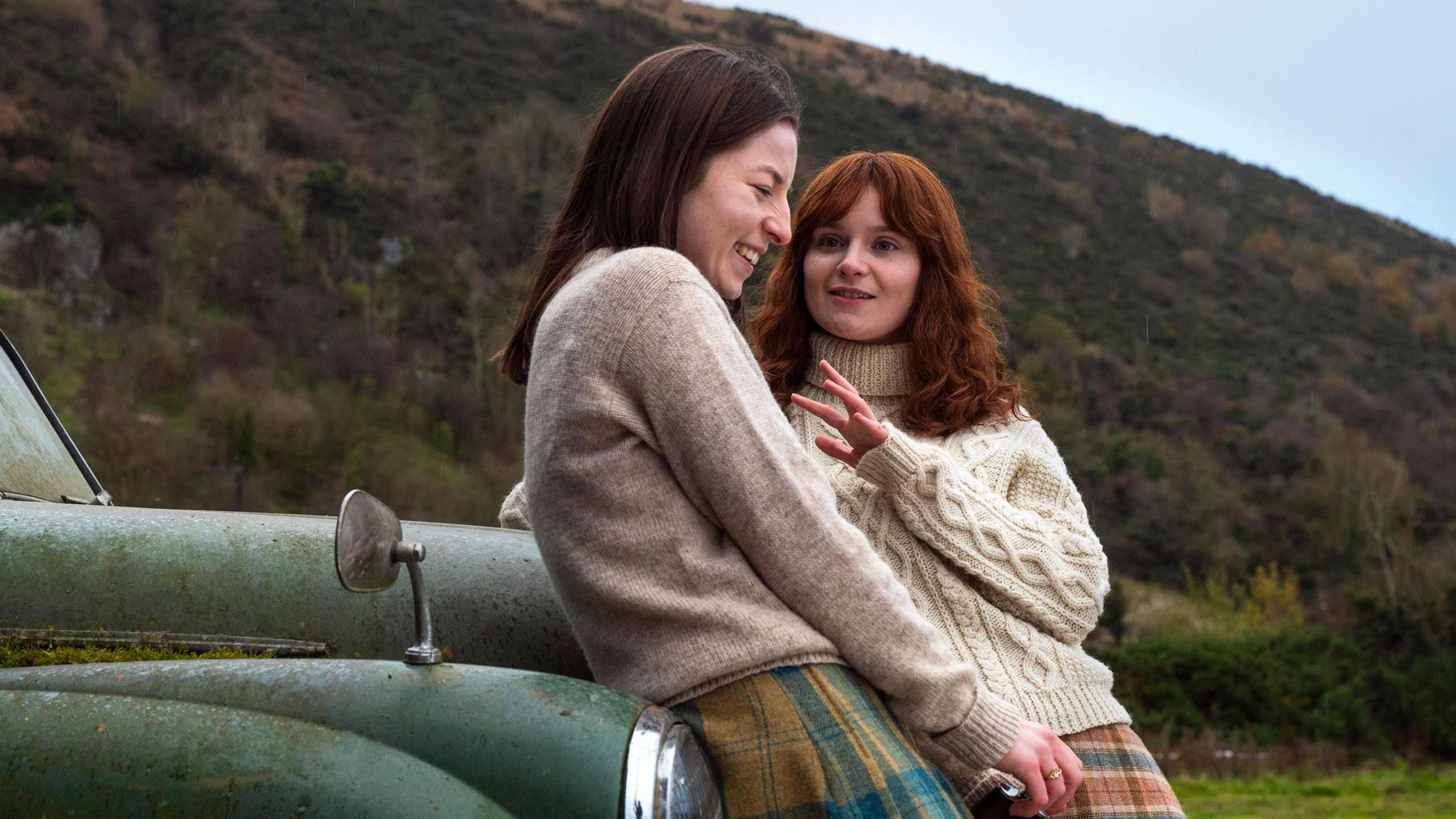 Hazel Doupe and Lola Petticrew in character, they are both leaning against an old green car, there is a mountain in the background. Hazel Doupe is wearing a cream jumper with a yellow and blue skirt. Lola Petticrew is wearing a light cream jumper with a red, yellow and blue skirt