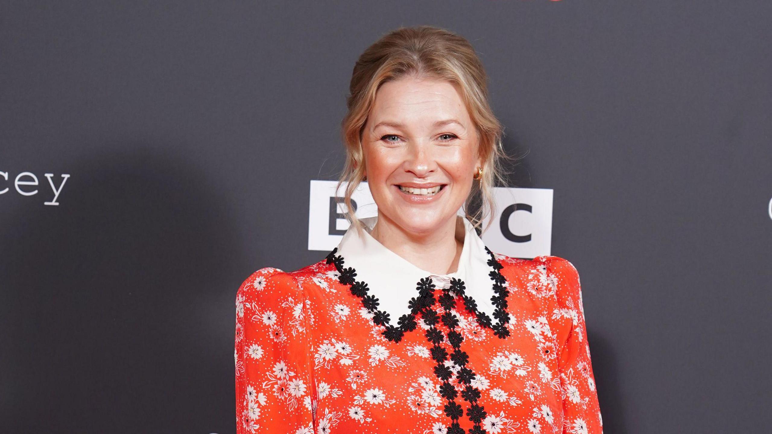 Joanna Page at launch event for Gavin and Stacey finale poses for photo in front of black background which says BBC behind her wears a red floral dress with large white collar and black floral embroidery around the collar and down the middle.