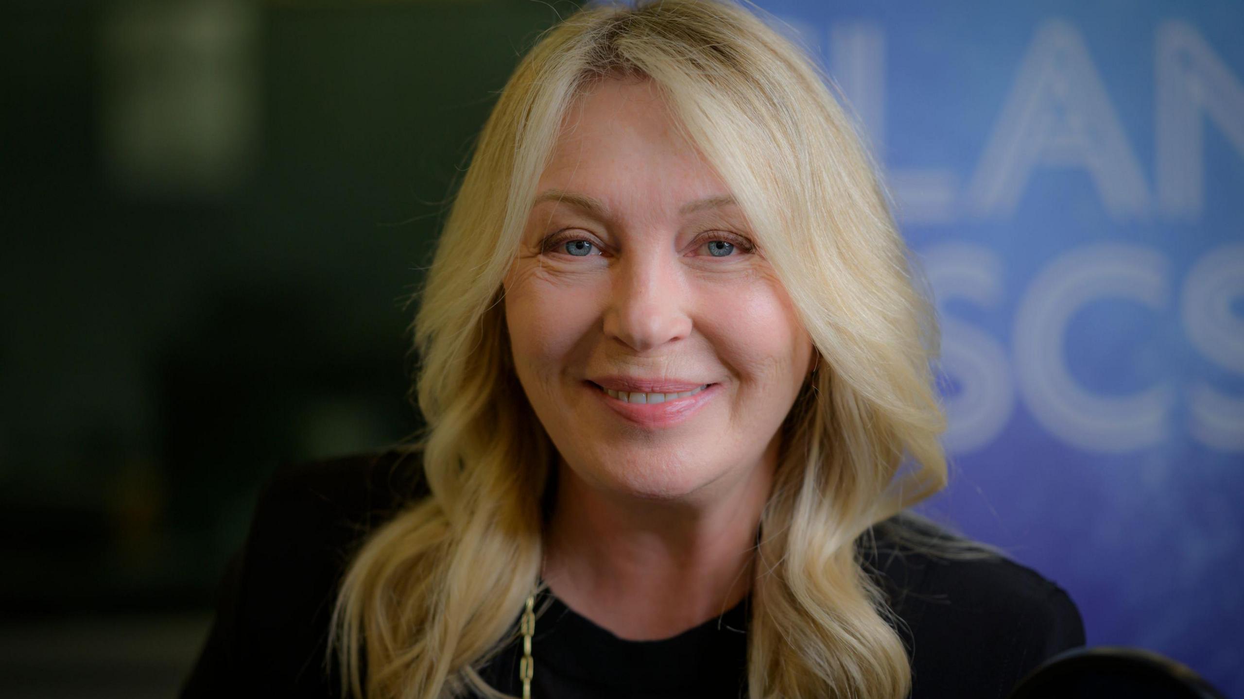 A woman, Kirsty Young, sits in a radio broadcasting studio. She has blonde wavy hair and is wearing a black blazer, black top and a gold necklace. She sits in behind a blue microphone with BBC Radio 4 Desert Island Discs written on it. 