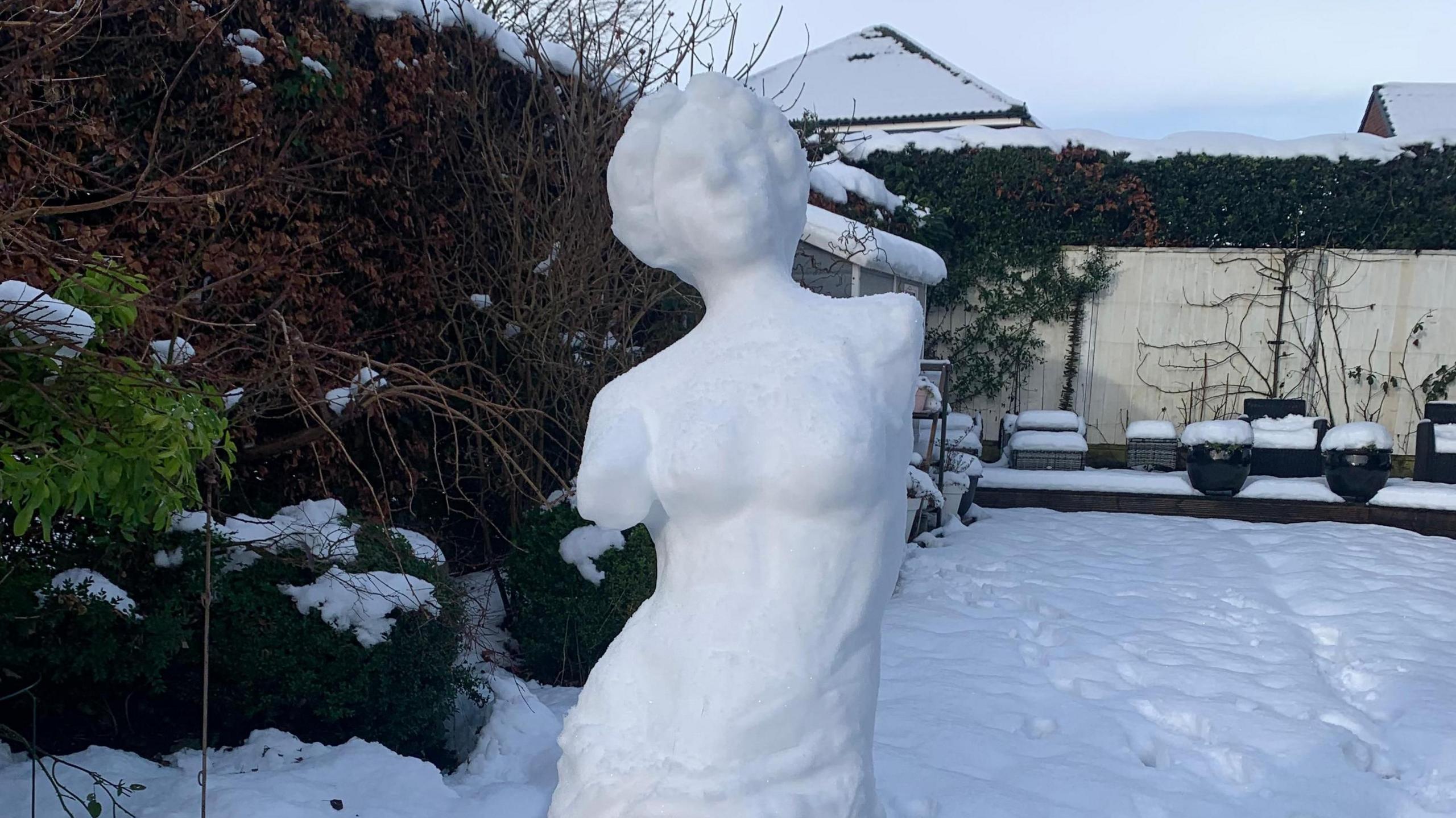 A classical Greek style sculpture of a woman with no arms made from snow. Behind the sculpture is a snowy suburban garden.