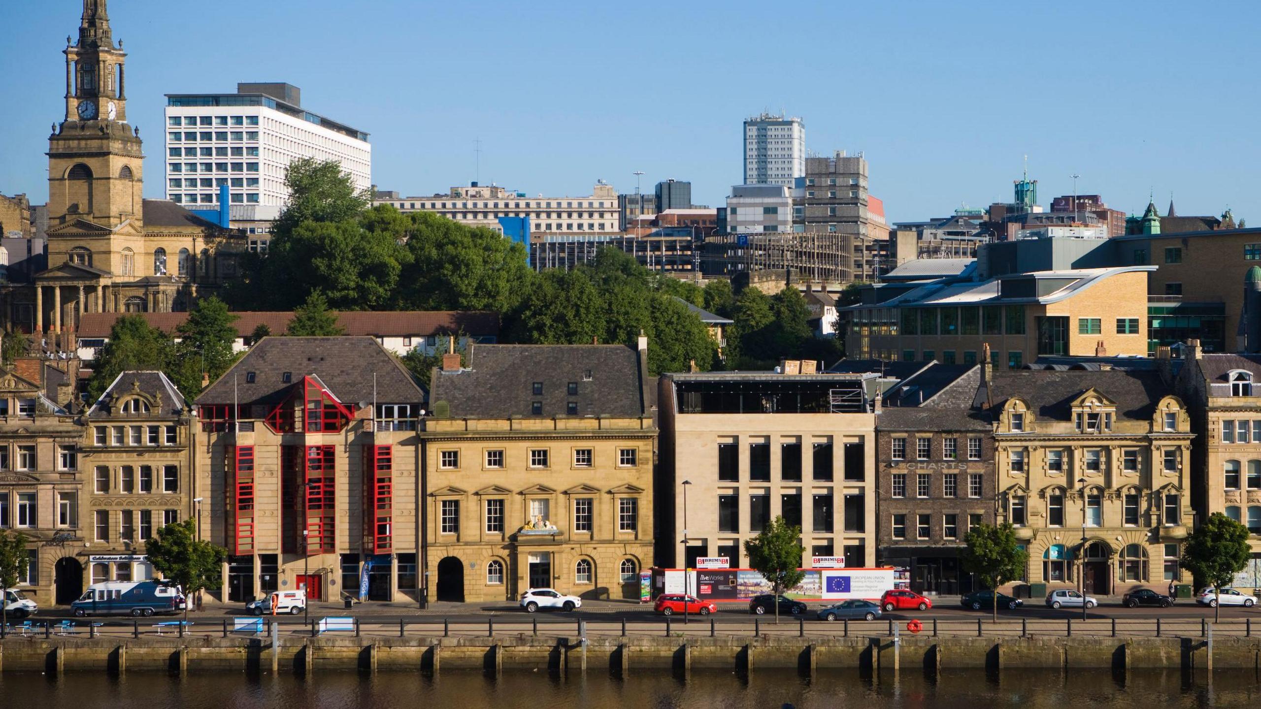 Multi-storey old-style light-coloured buildings with several windows along a riverfront with a church in the background.