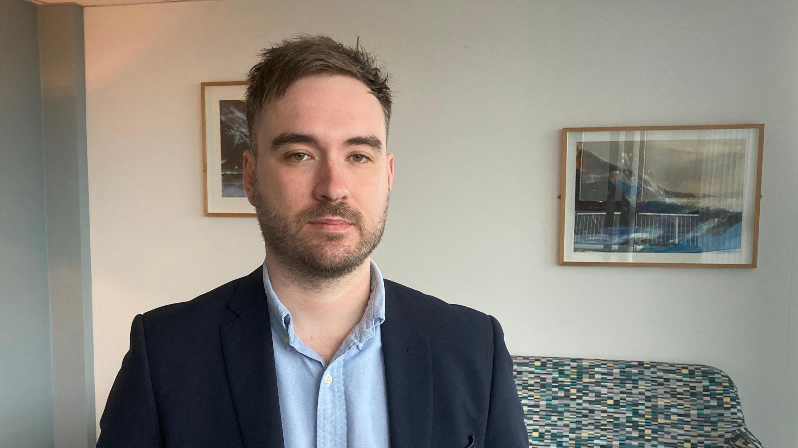 Dr Jamie Torrance is wearing a navy blue suit jacked and a light blue shirt with the top button undone. A multi-coloured sofa is visible behind him and there are framed photos of mountain landscapes on the wall 