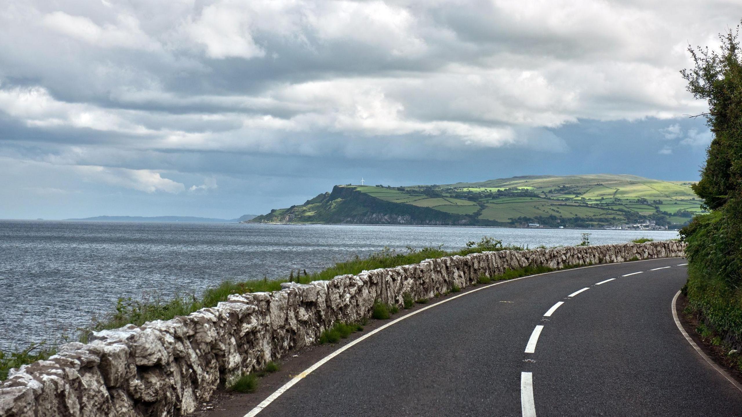 The coastal road to Carnlough, a village in County Antrim, Northern Ireland. A small stone wall separates the road form the sea, a cliff is seen in the distance