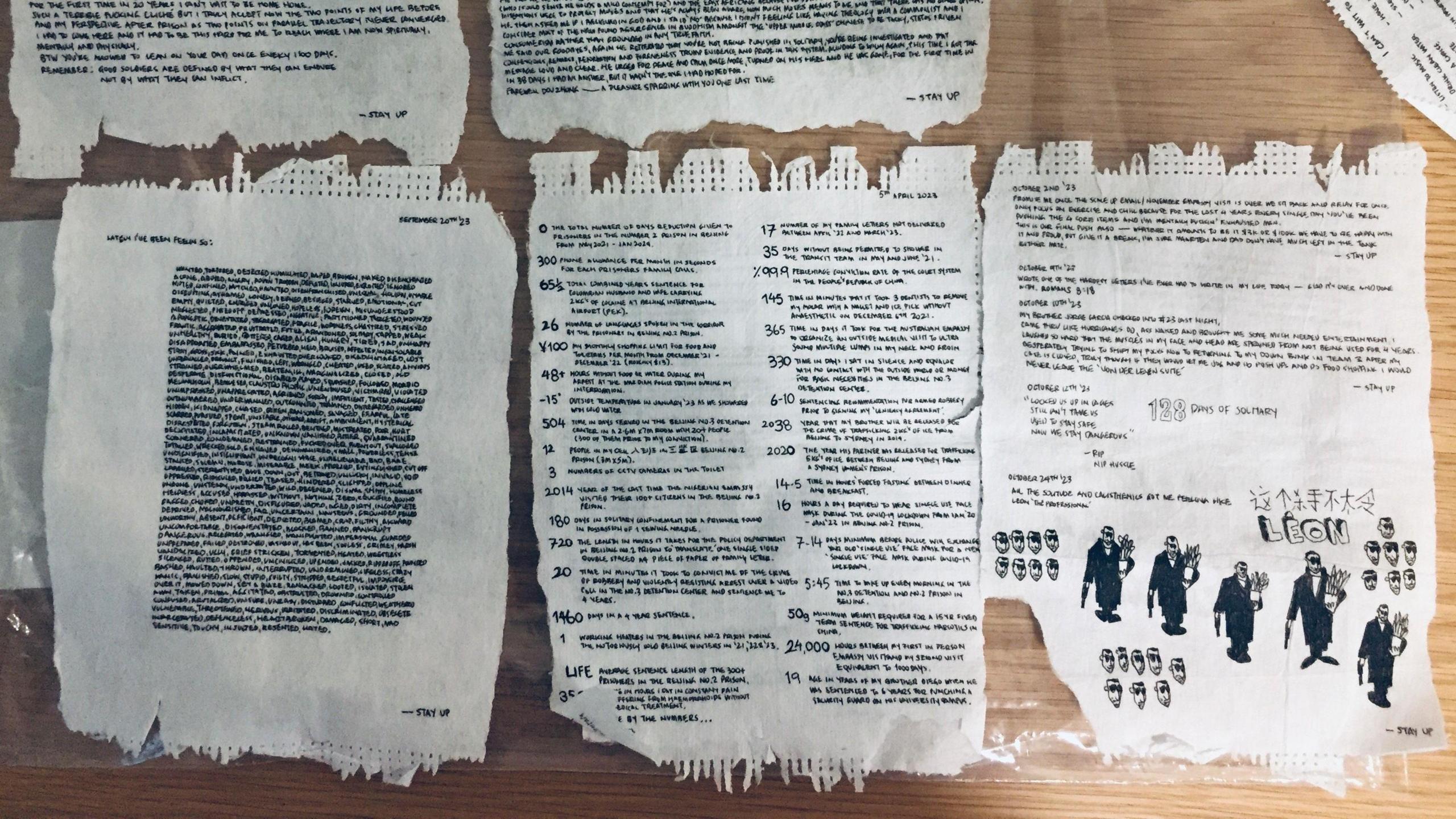Pages from Radalj's journal side by side on a wooden table. The pages have his writing and drawings., all in black pen