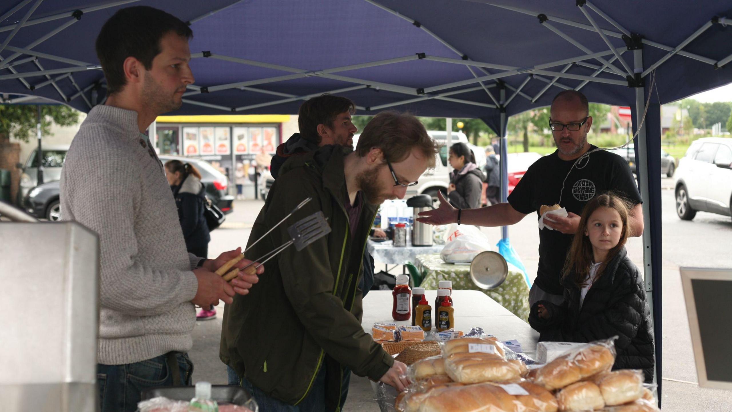 A group of people standing at a table with bread rolls and hot dog buns on it. There are also sauce bottles. One of the men is holding what looks like tongs and a spatula.