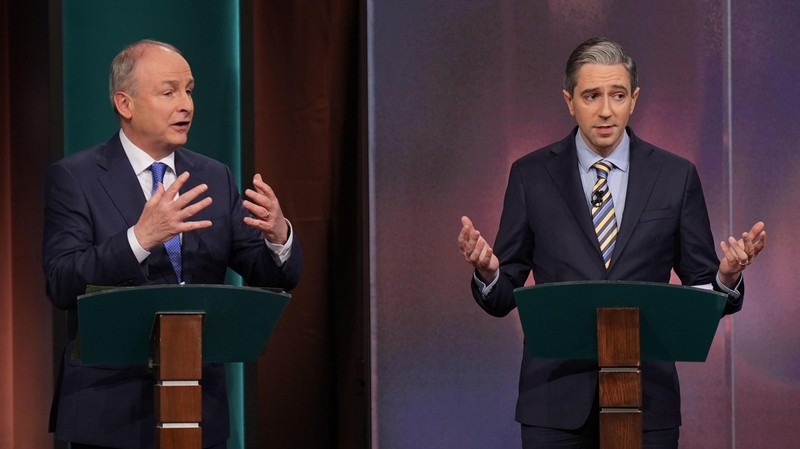 Micheál Martin and Simon Harris at the podium during a TV debate. Both men  are wearing dark suits and expressively using their hands 