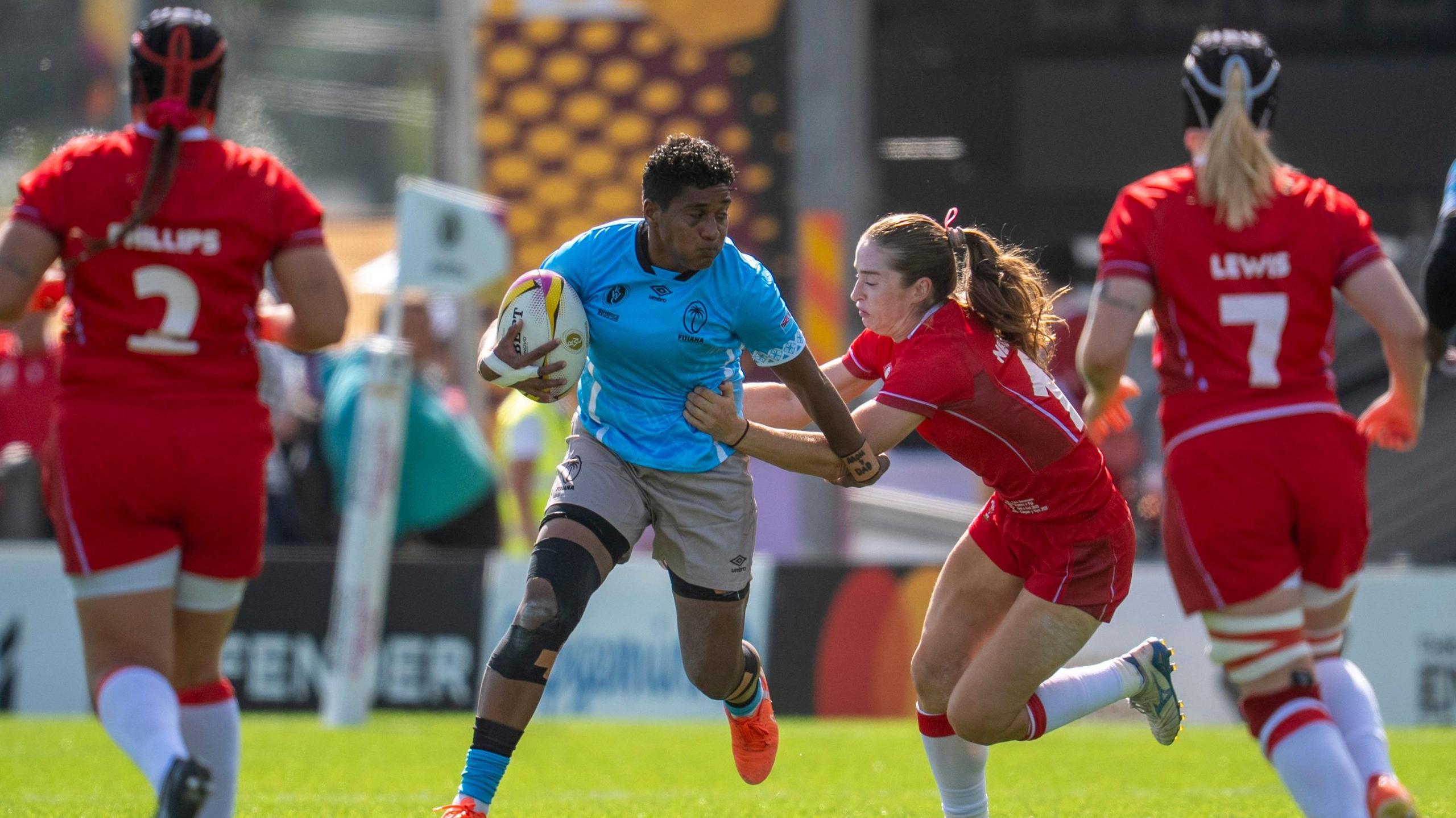 Fiji's Litiana Vueti and Wales' Lisa Neumann in action during the Women's Rugby World Cup 2025 pool B match at Sandy Park, Exeter. Two other Welsh players are running towards them. The Wales players are in red kits. The Fiji player is wearing a blue shirt with silver shorts.